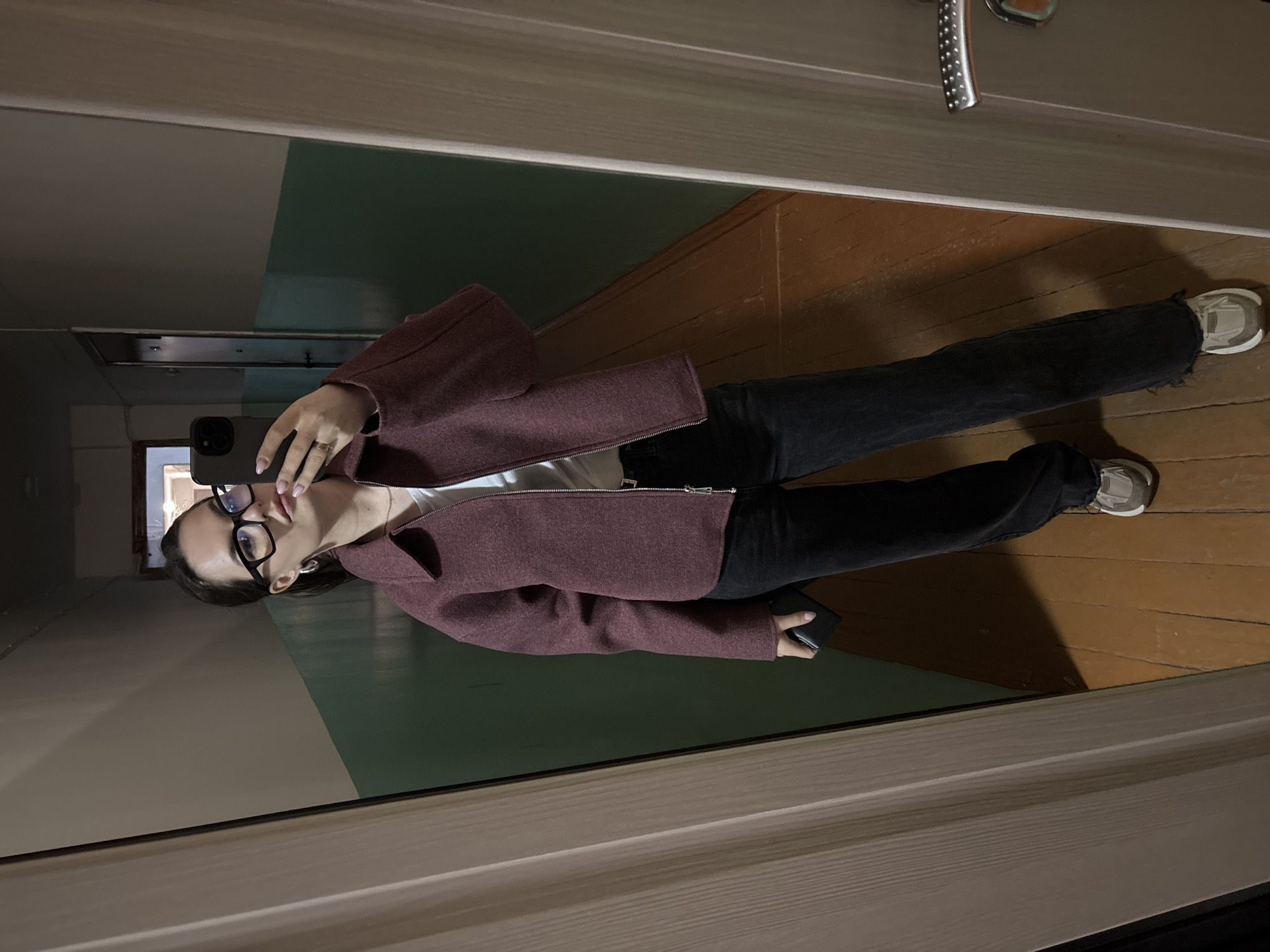 A woman is taking a selfie in a hallway. She is wearing glasses, a maroon jacket, black jeans, and sneakers. The hallway has wooden floors and painted walls with white on top and green on the bottom. A closed door is visible on the right side. The location is in the city of Chaikovsky, Russia.