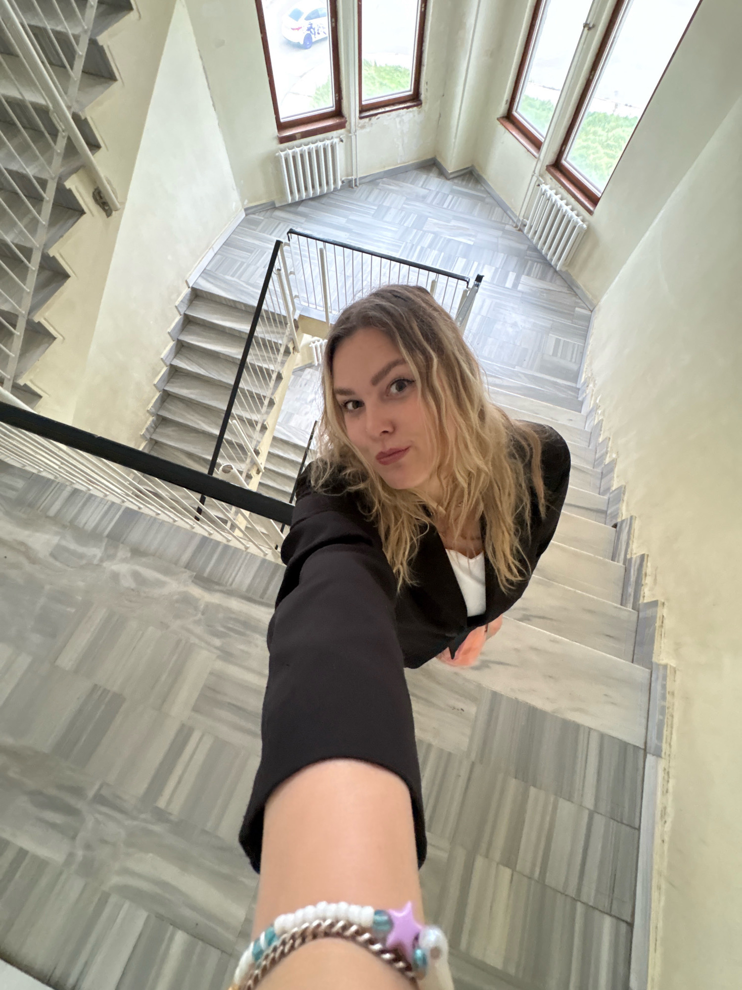 A woman with blonde, wavy hair is taking a selfie on a marble staircase. She is wearing a black blazer over a white top and a beaded bracelet on her left wrist. The staircase is wide with white railings and has a patterned marble floor. Windows are visible at the top of the staircase, overlooking an urban landscape that includes trees and a white car. The setting appears to be an interior staircase in a building. The location of Марковский, Russia is not discernible from the image.