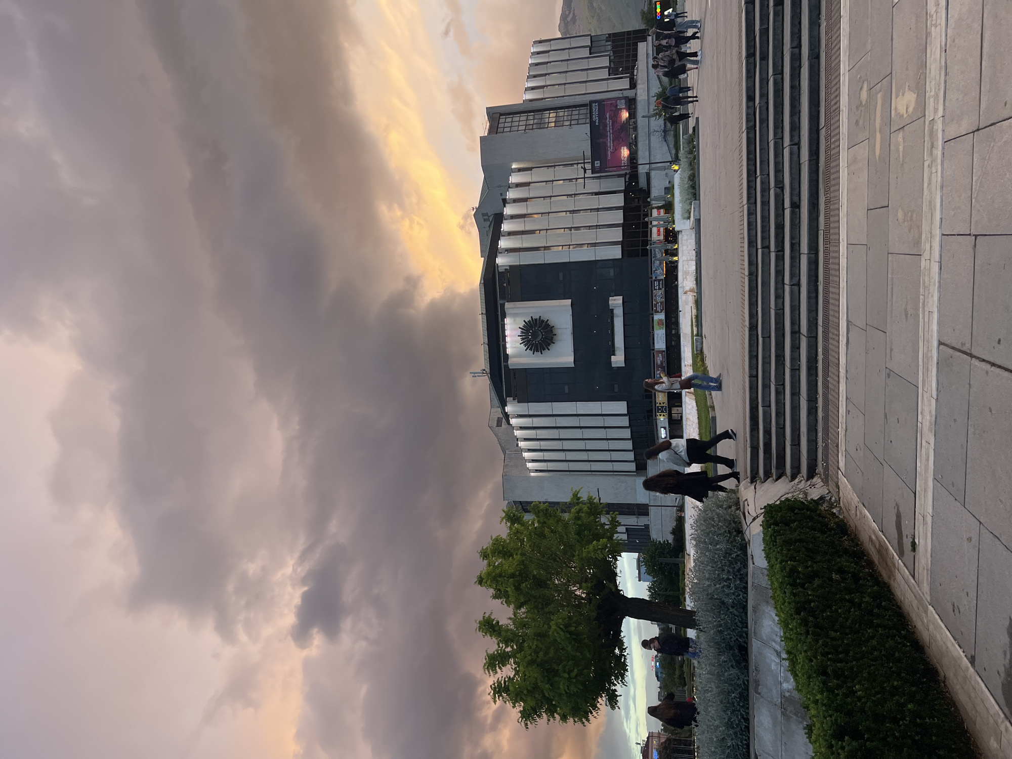 The image shows a view of the National Palace of Culture (NDK) in Sofia, Bulgaria, at sunset. A large, modern building is at the center, with a distinctive circular, sun-like decoration on its facade. A banner advertising a "Grande Partenza" event is visible on the side of the building. People are walking along a stone pathway and up a flight of concrete stairs adjacent to the building. A tree and trimmed bushes line the foreground. The sky is filled with dramatic clouds, displaying colors of grey, white, and a hint of yellow from the setting sun. A few individuals can be seen standing at the base of the building.