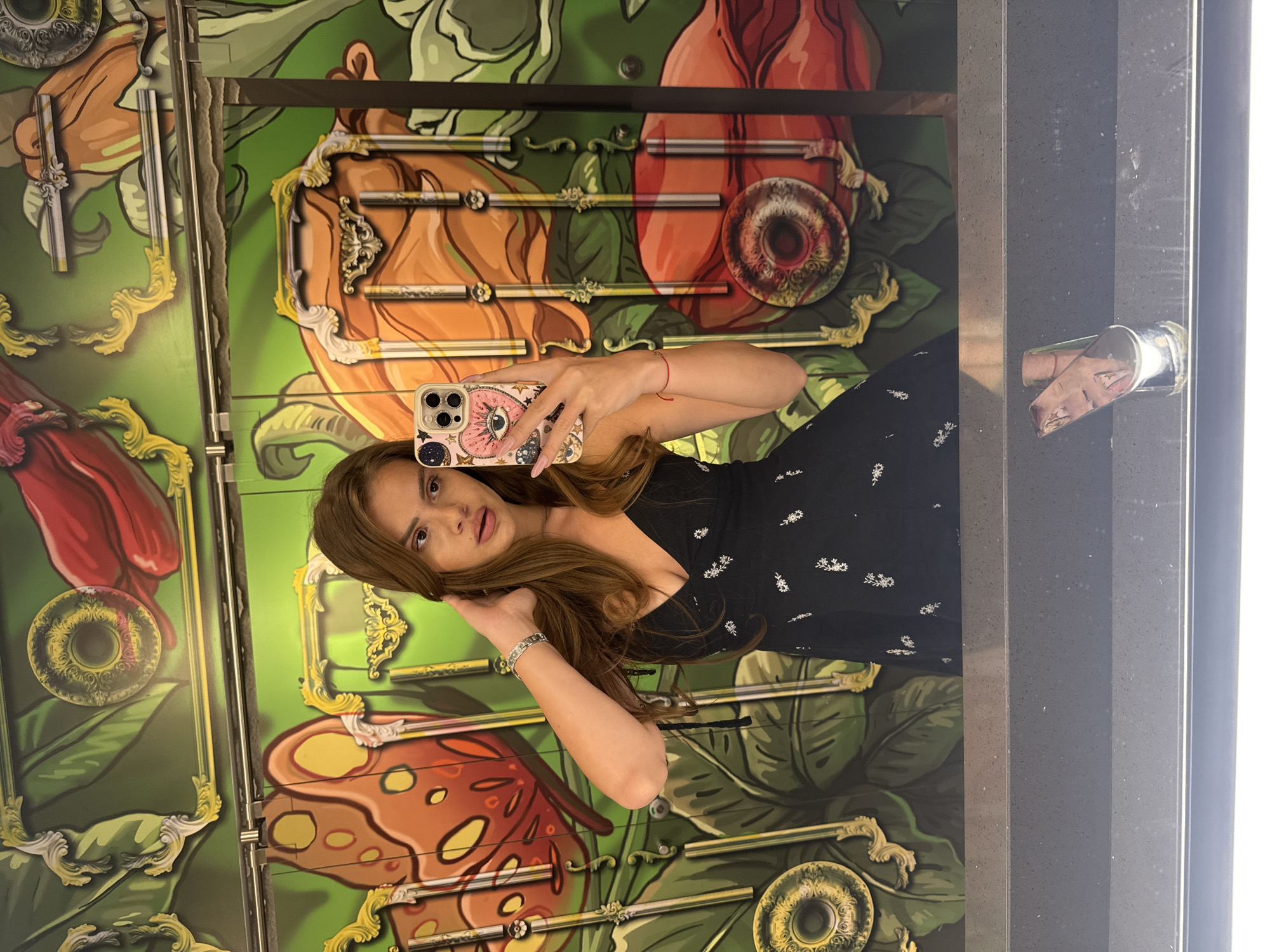 A young woman with brown hair is taking a mirror selfie in what appears to be a restroom. She is wearing a black dress with a floral pattern. The background is a vibrant mural depicting large, stylized flowers and butterflies, with ornate golden frames integrated into the design. The reflection in the mirror shows a countertop and a glass object.