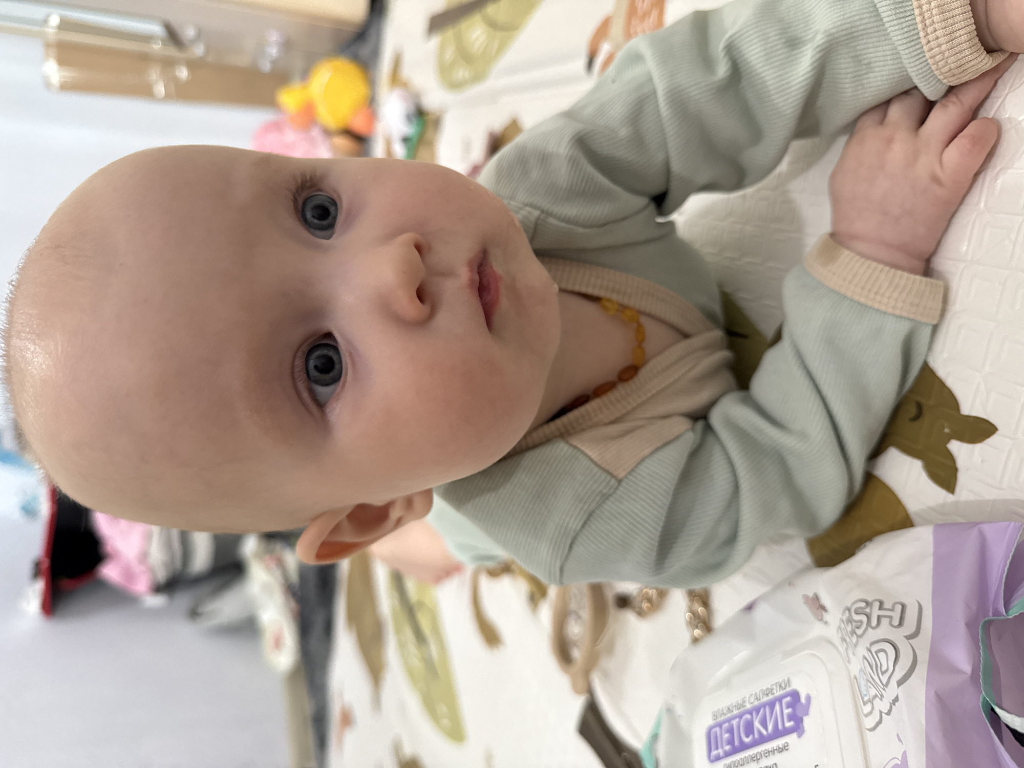 An image, originally rotated 90 degrees counter-clockwise, depicts an infant lying on their back and looking upwards at the camera. The baby has fair skin, prominent blue eyes, and very light, sparse hair. Their expression is calm and curious.
The baby is wearing a light mint-green long-sleeved top with contrasting beige ribbed cuffs and collar detailing, and an amber bead necklace is visible around their neck. They are positioned on a patterned surface, likely a playmat or blanket, featuring stylized natural motifs such as leaves and deer in shades of green, white, and brown.
In the blurred background, hints of other objects suggest an indoor setting, including a yellow toy and a pink item. A package of "FRESH LAND" baby wipes, with Cyrillic text reading "ВЛАЖНЫЕ САЛФЕТКИ" (Wet Wipes) and "ДЕТСКИЕ" (Children's) along with "гипоаллергенные" (hypoallergenic), is visible in the foreground. This Russian text provides a cultural cue consistent with the provided location context of Yakutsk, Russia.