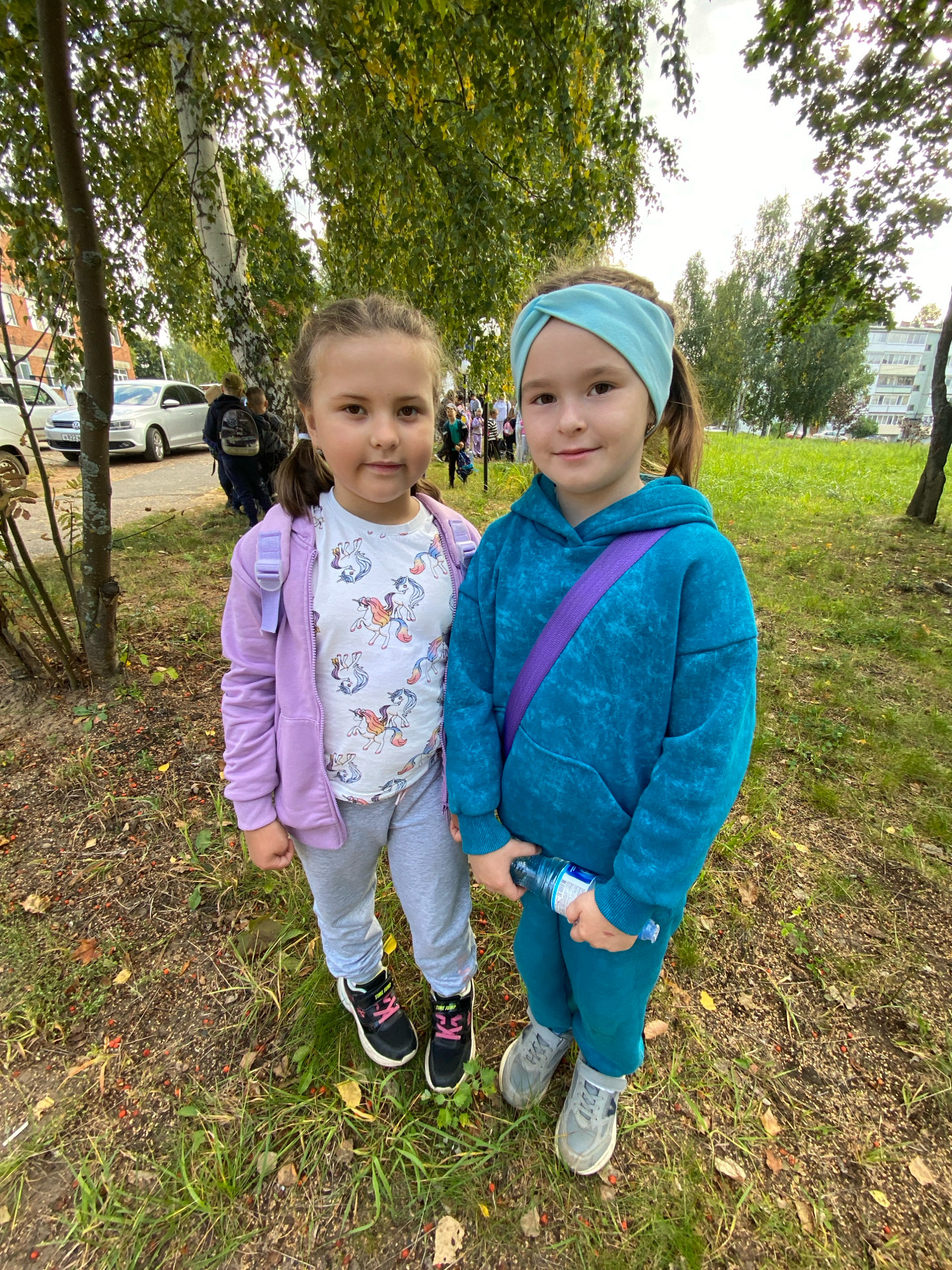 Two young girls stand side-by-side in an outdoor park-like setting with trees and grass. The girl on the left wears a lavender hooded jacket over a white t-shirt with a unicorn print and grey sweatpants. She has a small purple backpack on. The girl on the right wears a teal-colored hooded sweatshirt and matching teal pants, with a purple strap across her chest. She holds a clear plastic bottle of water. Both girls have their hair pulled back. In the background, a paved area with a white car and other people are visible, along with apartment buildings and more trees. The ground is covered with dry leaves and grass. The location is identified as Noviy, Russia.