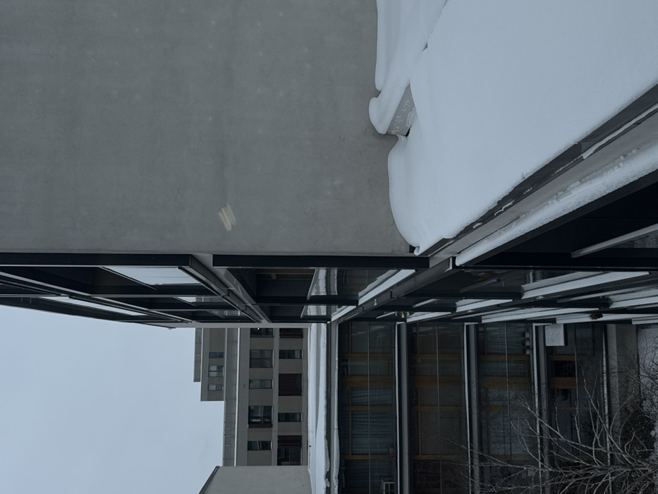 This image presents a forensic view of a modern residential complex in Warsaw, Poland, under overcast winter conditions. The scene is dominated by contemporary multi-story apartment buildings constructed with light gray, textured facades, likely concrete, and features dark-framed windows and balconies.
In the immediate foreground and to the right, a large section of a building's gray wall is visible, marked by two faint, light-colored horizontal streaks. A significant accumulation of snow covers a flat ledge or balcony railing extending from this building in the lower right. To the left, a series of identical dark-framed windows and glass-paneled balconies are stacked vertically across multiple floors, reflecting the sky and adjacent structures. Many of these windows exhibit drawn interior curtains.
Further into the mid-ground and background, additional geometric apartment buildings with similar gray facades and dark-framed windows are visible, appearing terraced or stepped. All exterior horizontal surfaces, including roofs, ledges, and balcony floors, are uniformly covered with fresh snow, indicating a recent snowfall. A single bare deciduous tree, lightly dusted with snow on its branches, occupies the lower-left quadrant of the image. The sky is a uniform, overcast gray, casting diffused light across the scene. No individuals, vehicles, or explicit activities are visible.