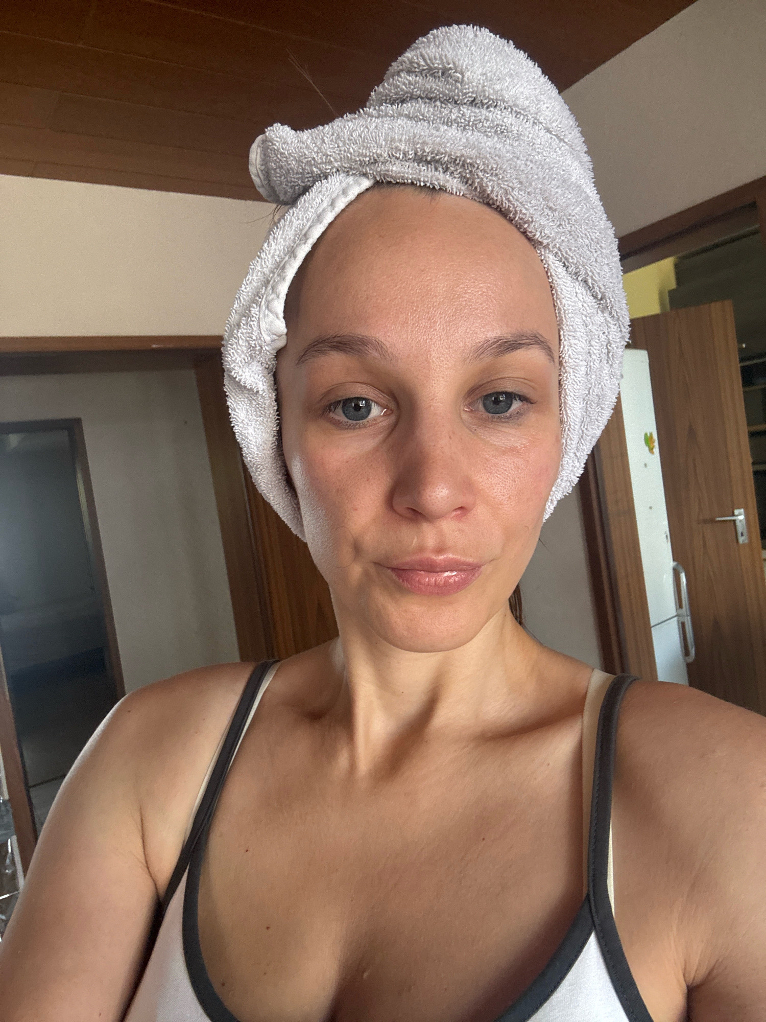 A woman with a gray towel wrapped around her head is looking directly at the camera. She is wearing a white tank top with gray trim. The background shows a wooden door and a wall. The lighting suggests it is daytime.