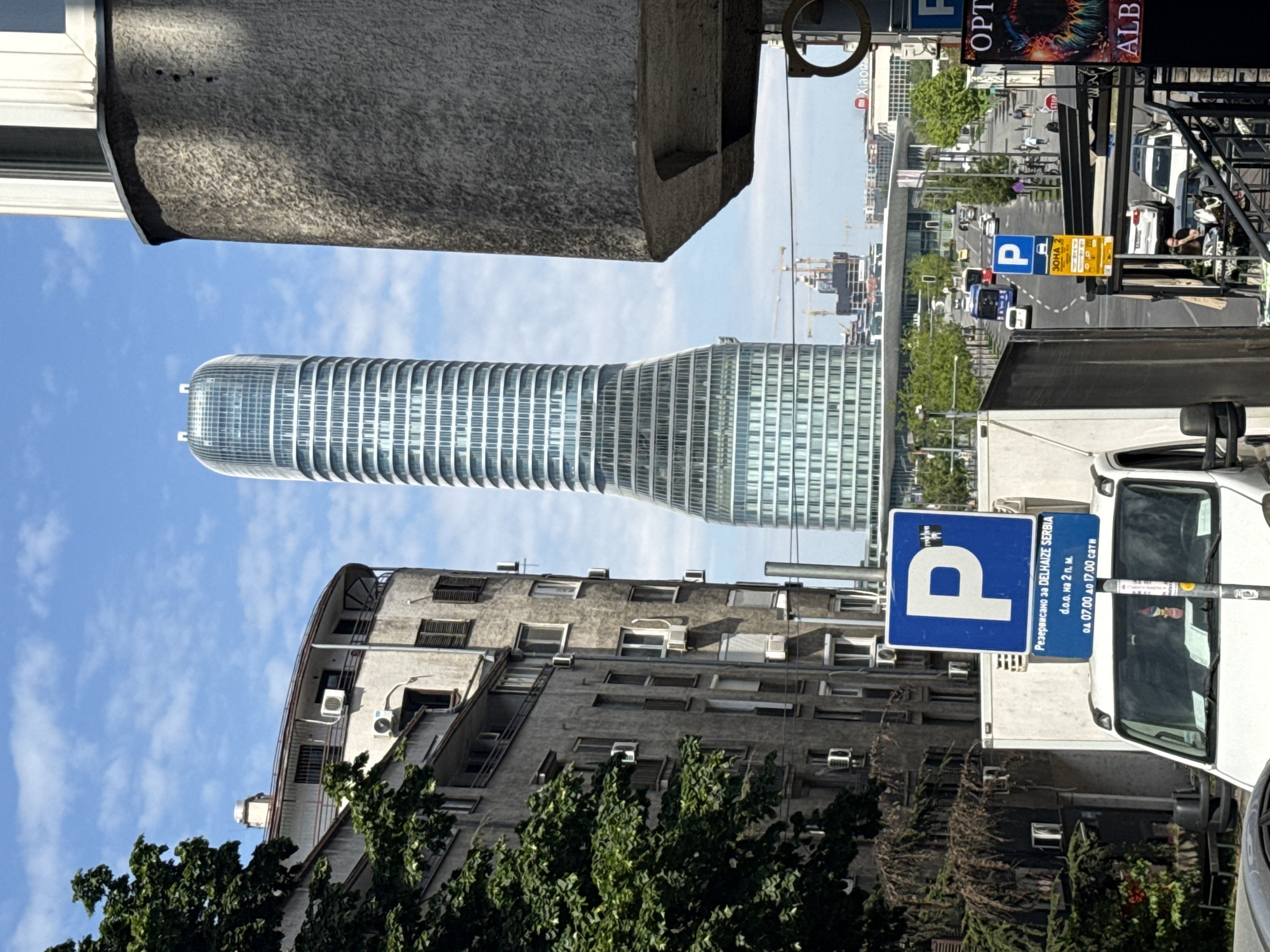 The image, taken in Belgrade, Serbia, shows a street scene featuring a modern, cylindrical skyscraper in the background. The perspective is somewhat skewed, taken from a low angle, looking up. In the foreground, there are details of an older building and a parking sign. The parking sign is blue and white with a large "P" on it. It also has text in Serbian. The sky is visible with scattered clouds, providing a bright backdrop. Street signs and traffic are visible in the mid ground, suggesting a busy urban area. The image is composed of old and new architecture.