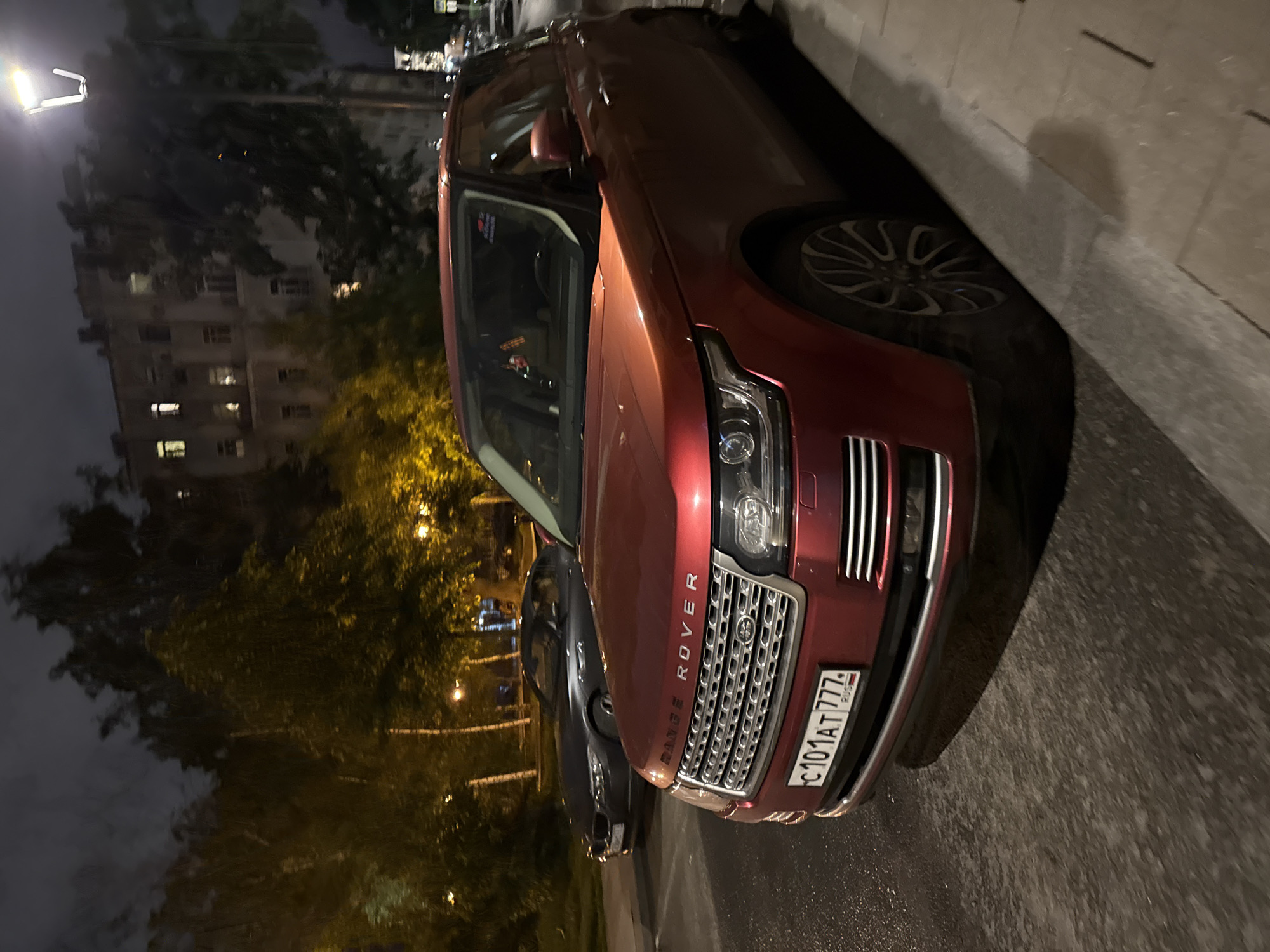 A red Range Rover is parked on a street in Moscow, Russia. The car has the license plate "C101AT 777". The street is paved, and there is a curb next to the car. Behind the car are buildings and trees. A streetlamp is visible in the upper left corner of the image. It is night, and the scene is lit by streetlights. A gray car is partially visible behind the Range Rover.