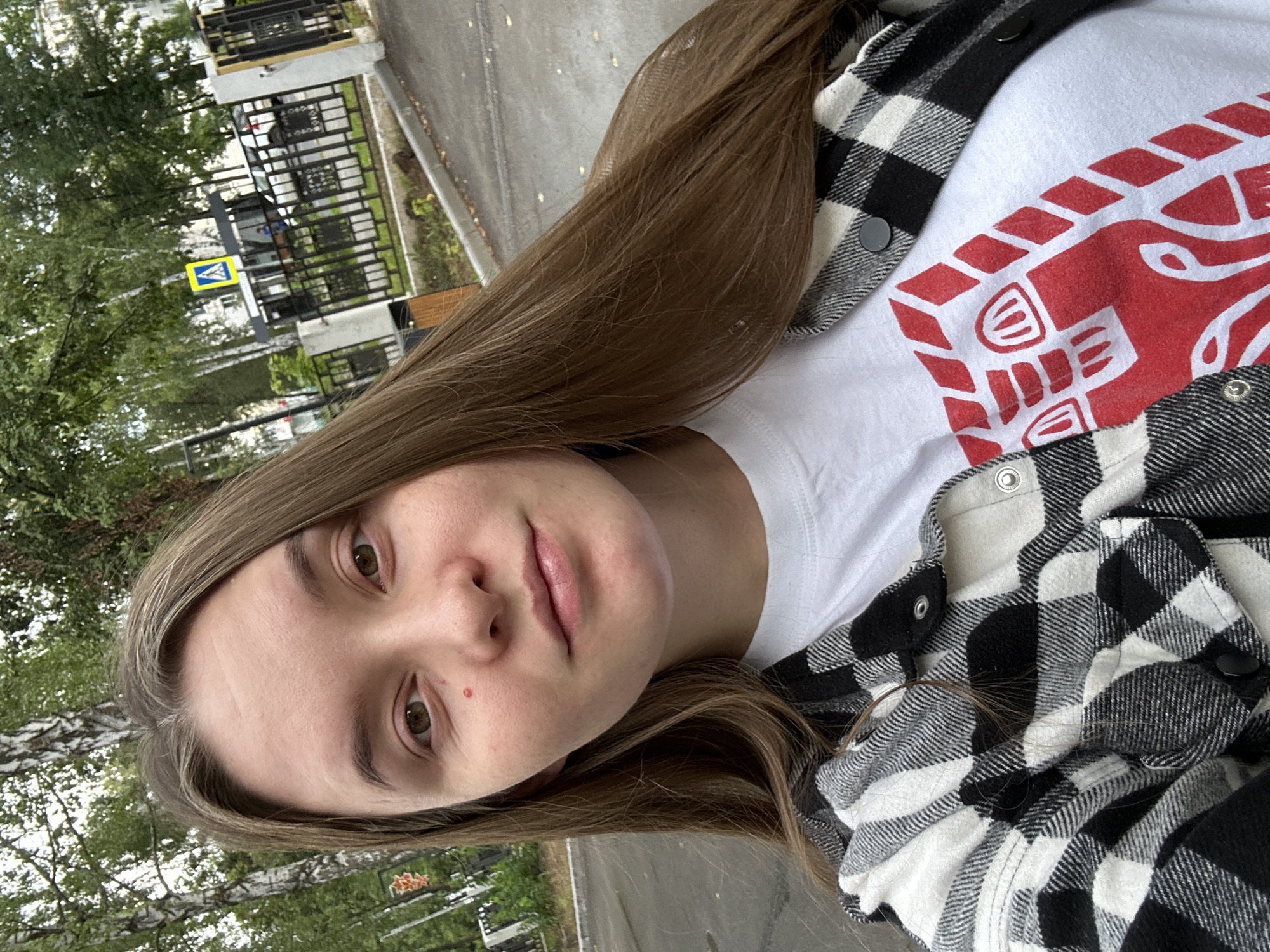 The image is a close-up, selfie-style photograph of a young woman with long, brown hair, wearing a white t-shirt with a red and white graphic design and an open black and white plaid shirt over it. She is outdoors, with trees and a fence visible in the background. A street sign with a pedestrian crossing symbol is also present. The overall scene suggests a casual outdoor setting, likely in a residential or park area of Chaikovsky, Russia, given the commonality of such natural and urban elements in Russian cities.