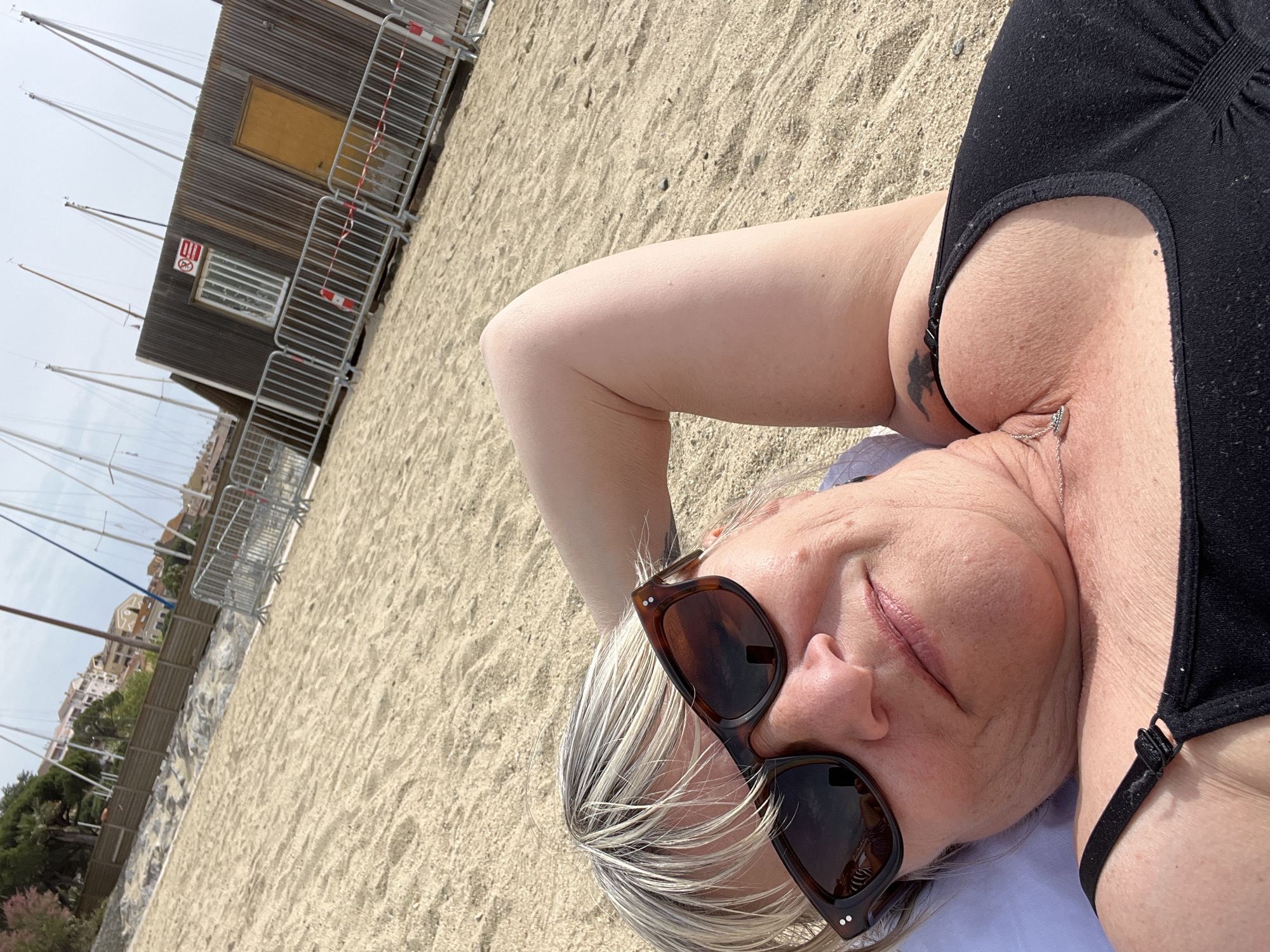 A woman lies on a sandy beach, presumably near Sainte-Maxime, France, based on the presence of buildings and sailboat masts in the background. She is wearing a black one-piece swimsuit and sunglasses with dark lenses and tortoiseshell frames. Her hair is blonde with graying roots. A small building with metal fencing and a warning sign is visible in the background, along with a row of sailboat masts. The environment suggests an outdoor location during daylight.