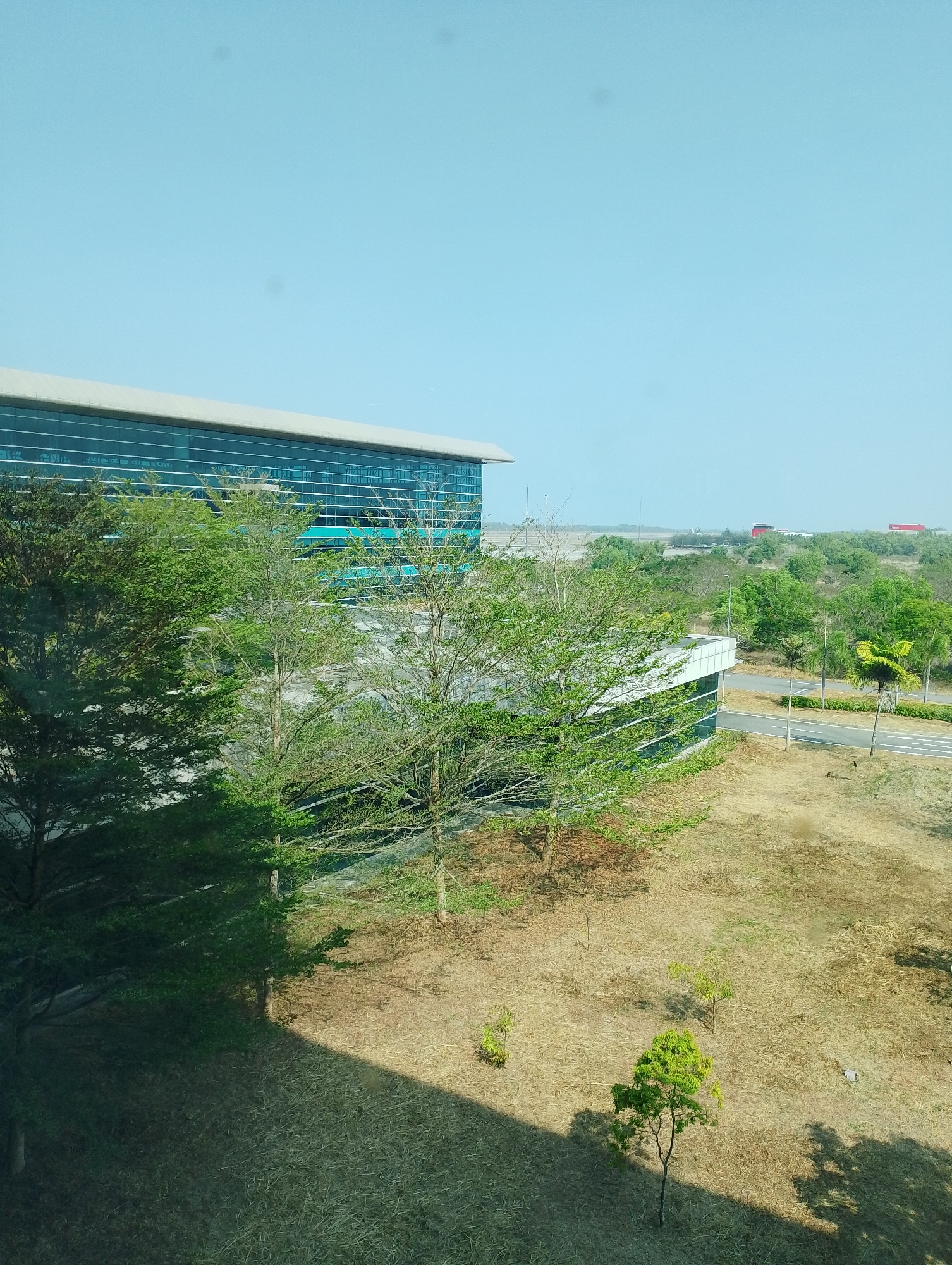 The image shows a view of the interior of a building complex in Palihan, Indonesia, featuring modern architecture with glass facades and a large open space. The building, which appears to be an airport terminal, has a long, horizontal structure with a light-colored roof and blue-tinted glass windows reflecting the sky. In front of the building, there are several trees with sparse green foliage, casting shadows on the dry, tan-colored ground. A paved road runs alongside the building, with streetlights visible. In the distance, the image shows more trees, a flat landscape, and what appears to be an airport runway with some distant red structures. The sky is a clear, light blue, indicating a sunny day.