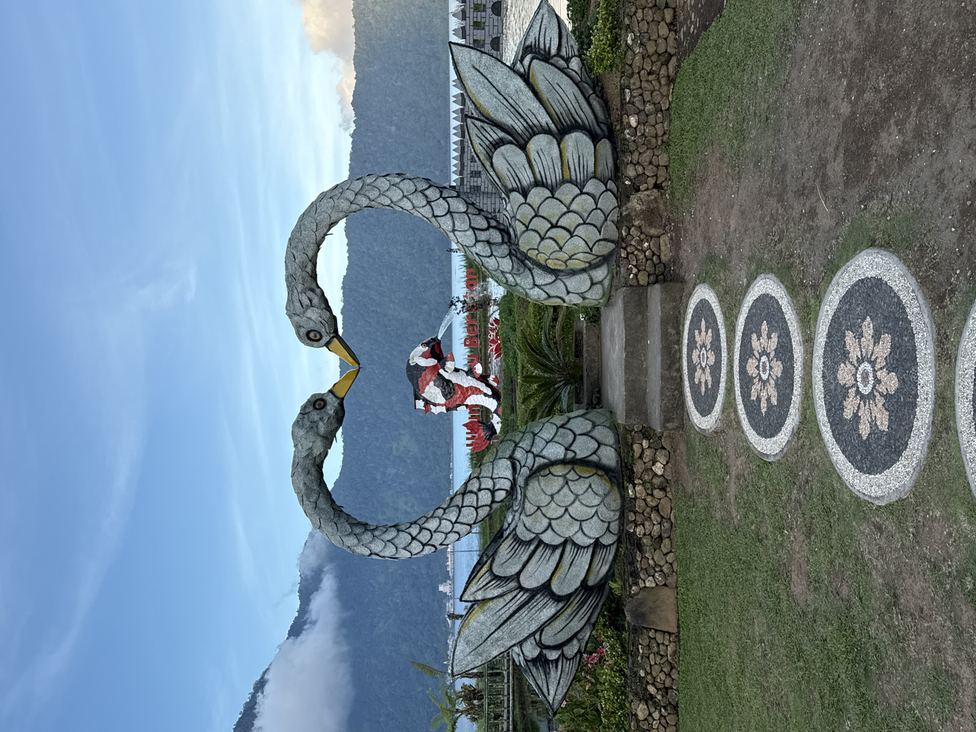 The image shows a swan statue in Baturiti, Indonesia. The statue consists of two gray swans with yellow beaks, their necks curved to form a heart shape. Behind the swans, there is a body of water with mountains in the background. The sky is blue with some clouds. In front of the statue, there is a path with circular stone designs. The scene is set in a garden environment with grass and stone borders.