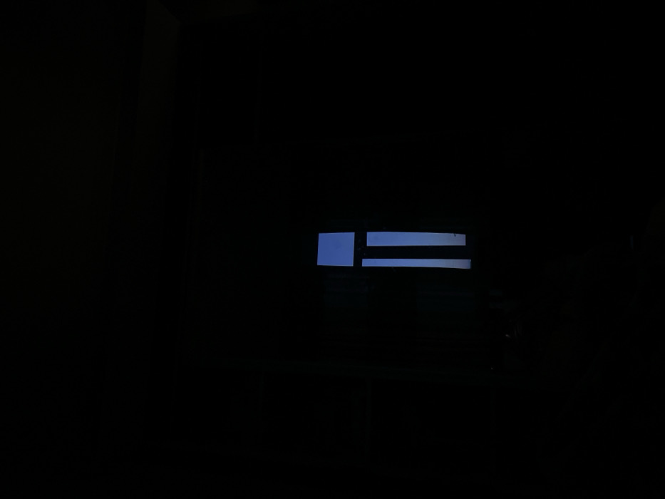 This image depicts an interior space in Navoiy City, Uzbekistan, characterized by profound darkness. A rectangular window is positioned on the right half of the frame, serving as the sole source of illumination. Through this window, a clear, solid blue sky is visible, suggesting daylight conditions outside. The window structure includes two distinct vertical dark elements, consistent with window frames or mullions, dividing the visible sky into three vertical sections. The surrounding interior walls, floor, and ceiling are entirely obscured by deep shadow, rendering any additional subjects or objects undetectable. No individuals or specific actions are discernible within the scene.