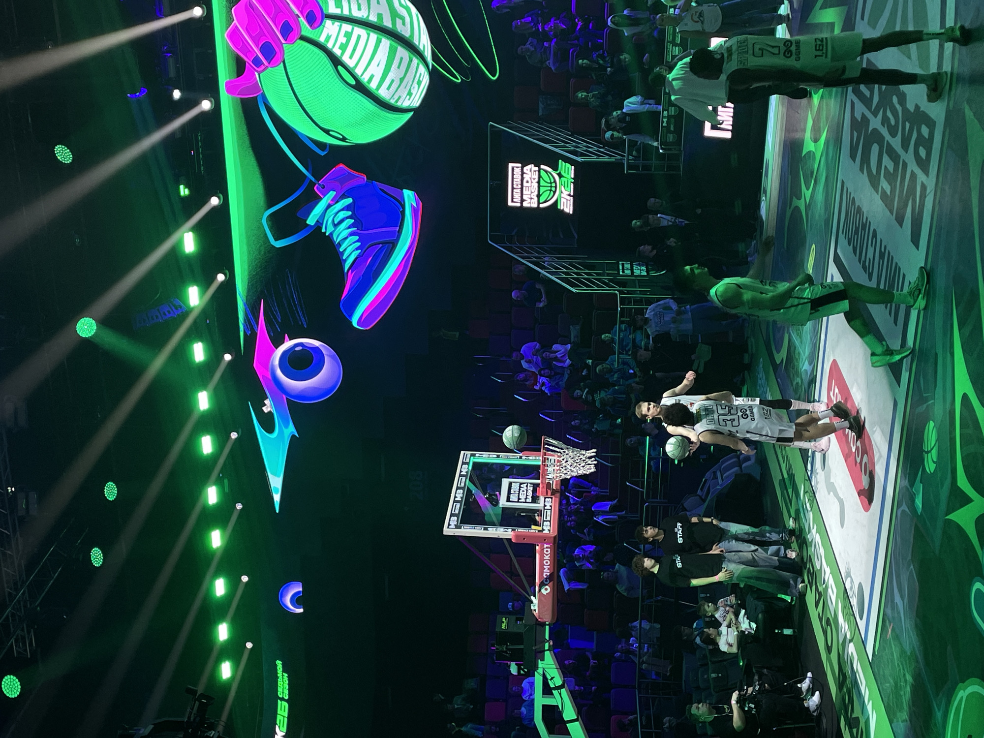 The image shows a basketball game or event, possibly related to "Liga Stavok Media Basket", based on the signage. The event appears to be in a dark, illuminated venue with bright green and purple lighting and projection effects, including stylized graphics of a basketball, shoe, and an eye. There are spectators in the background, and basketball players are visible on the court. A basketball hoop is present, and the words "MEDIA BASKET" are prominently displayed on the court surface. There are staff members standing near the court. The Russian words "ЛИГА СТАВОК СИЛЬНОЙ СЕЗОН" (Liga Stavok Strong Season) can be seen. The scene suggests a live basketball event with a focus on media presence and promotion. It is implied from other visible elements that this is occurring in Moscow, Russia.