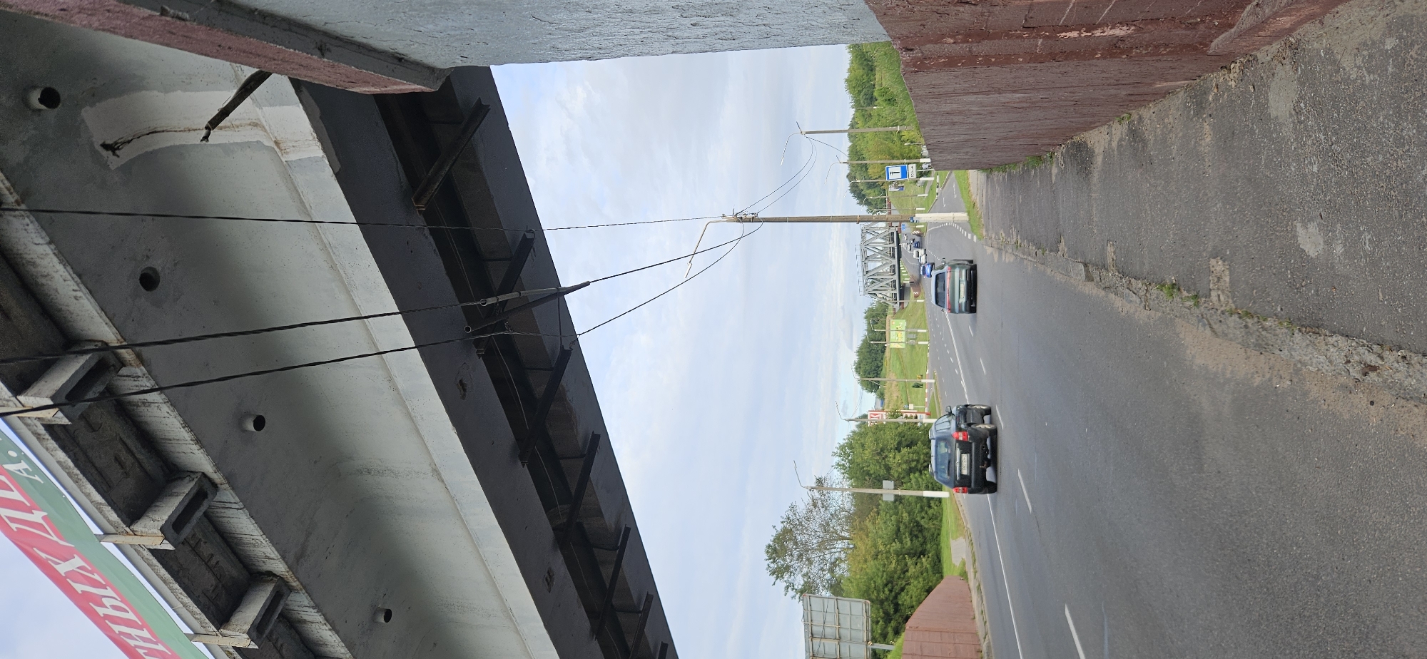 The image shows a street scene in Vitsebsk, Belarus, looking towards a railway bridge in the distance. Overhead, a concrete overpass dominates the foreground, with power lines suspended from it. Below the overpass, a road stretches into the distance, with several cars visible. A dark-colored SUV is in the left lane, followed by a lighter-colored hatchback in the right lane. Further down the road, more vehicles are visible, and a large metal railway bridge spans the horizon. To the right of the road, a concrete sidewalk runs alongside a red-brown wall. The sky is overcast with scattered clouds, and lush green trees line the sides of the road. The overall impression is one of a typical urban streetscape on a cloudy day, with the prominent infrastructure of the overpass and railway bridge suggesting an area with significant transportation networks.
