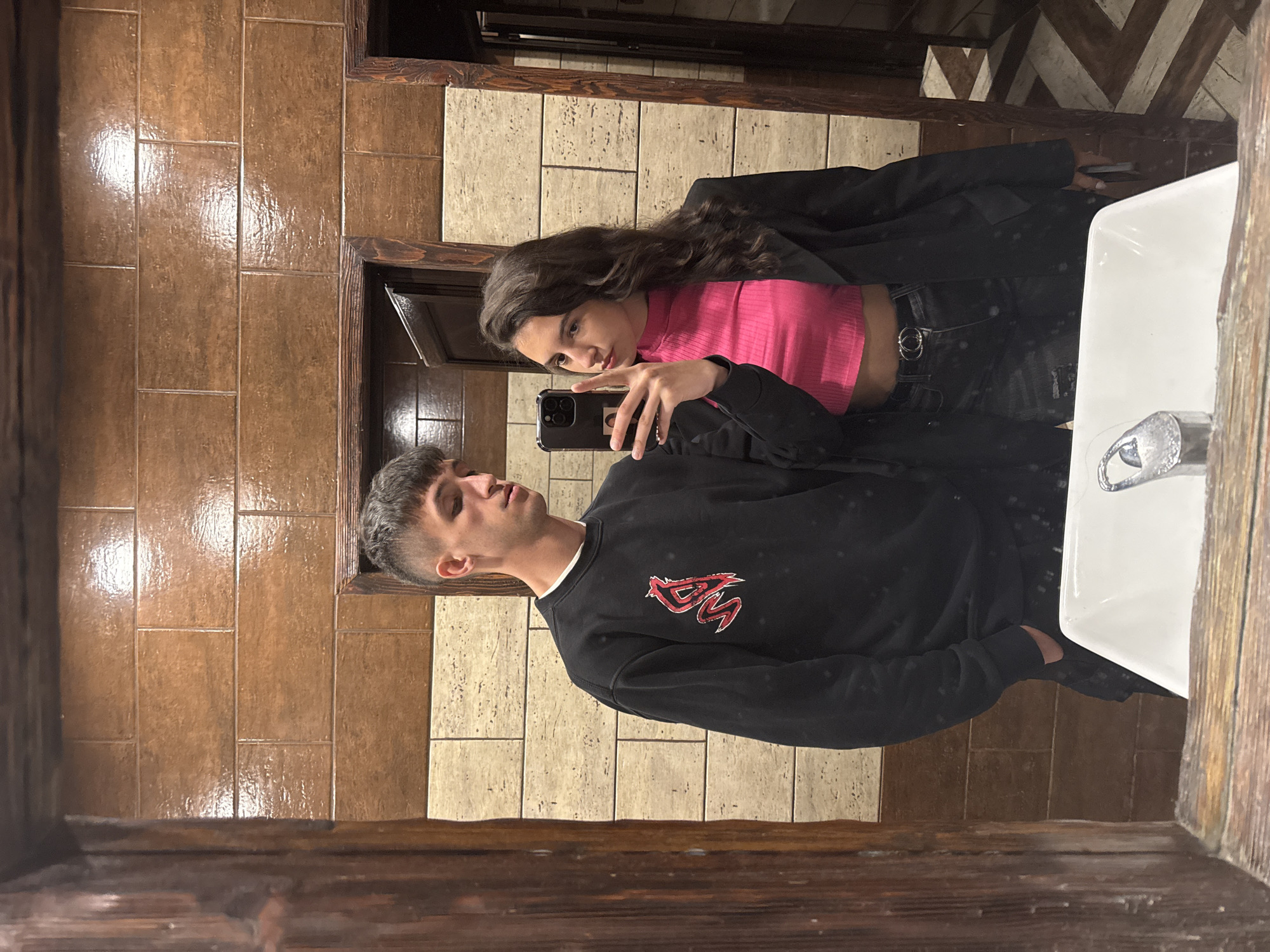 A young man and woman are taking a selfie in a bathroom. The bathroom has brown and beige tile walls and a dark wood-framed mirror. The woman is wearing a pink top and a black jacket. The man is wearing a black sweatshirt with a red graphic. The woman is holding a black smartphone to take the photo. The location may be Stara Zagora, Bulgaria.