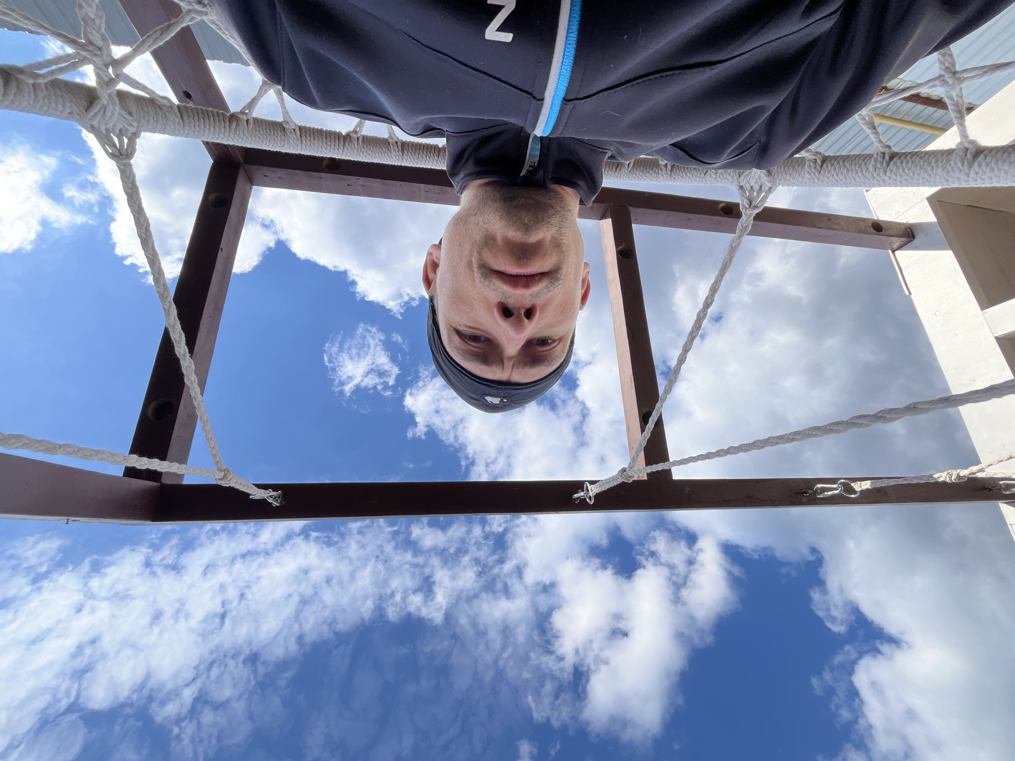 The image, taken in Chaikovsky, Russia, presents a unique low-angle view of a man looking directly at the camera while appearing to be upside down. The camera is positioned beneath him, looking upwards through the structure he is in.
The central subject is a man's face, framed by a dark bandana or hair, with a neutral to slightly serious expression and a direct gaze. He is wearing a dark, long-sleeved athletic jacket with a blue and white zipper accent on the front. A prominent white letter 'Z' is visible on the left side of his chest.
Around the man, the white, braided ropes of a hammock's netting form a crisscross pattern. These ropes are attached to a sturdy brown wooden frame, which outlines the upper part of the image. The arrangement suggests the man is lying in a hammock or a similar suspended rope structure, with the camera pointing up from below.
The background consists of a bright, clear blue sky adorned with scattered white, fluffy clouds, indicating a sunny day. The overall impression is one of leisure or a playful perspective in an outdoor setting. The bright lighting suggests it is daytime with fair weather.