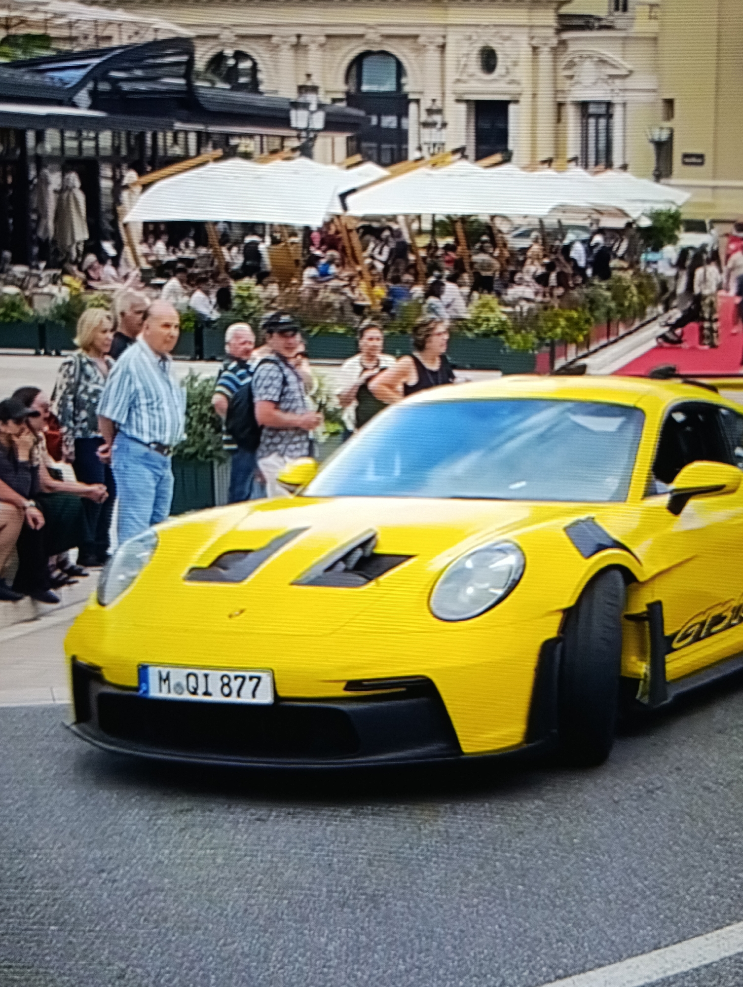 The image shows a yellow Porsche GT3 RS parked on an asphalt street. Behind the car, a group of people are standing and sitting, observing. Further back, there's an outdoor seating area with white umbrellas and numerous people, suggesting a restaurant or cafe. The architecture in the background features a light-colored building with arched windows and a dark awning or structure. The scene appears to be in a European city, based on the architecture and vehicle license plate. The specific city is not possible to determine.