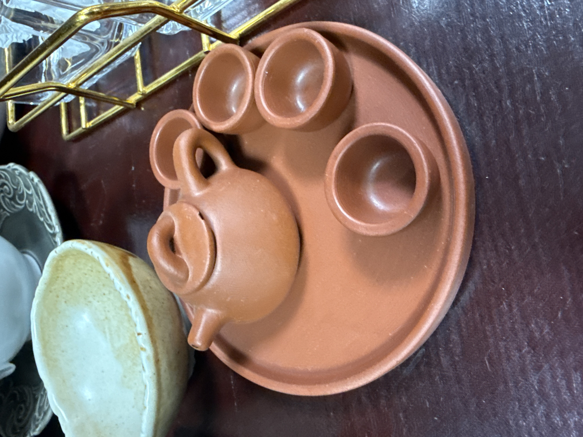 A high-angle, close-up shot shows a small, terracotta-colored tea set resting on a dark, textured surface, possibly a wooden table. The set includes a round tray with indents to hold three small teacups and a teapot with a rounded body and a spout. Another teacup, cream-colored with brown edges, is visible on the left side of the image, along with fragments of a gray plate with a swirling pattern and a shiny, clear glass object with a gold wire frame. The location cannot be definitively determined as Ust-Koksa, Russia, or any other specific location, as there is no visual information to connect the scene to any place.