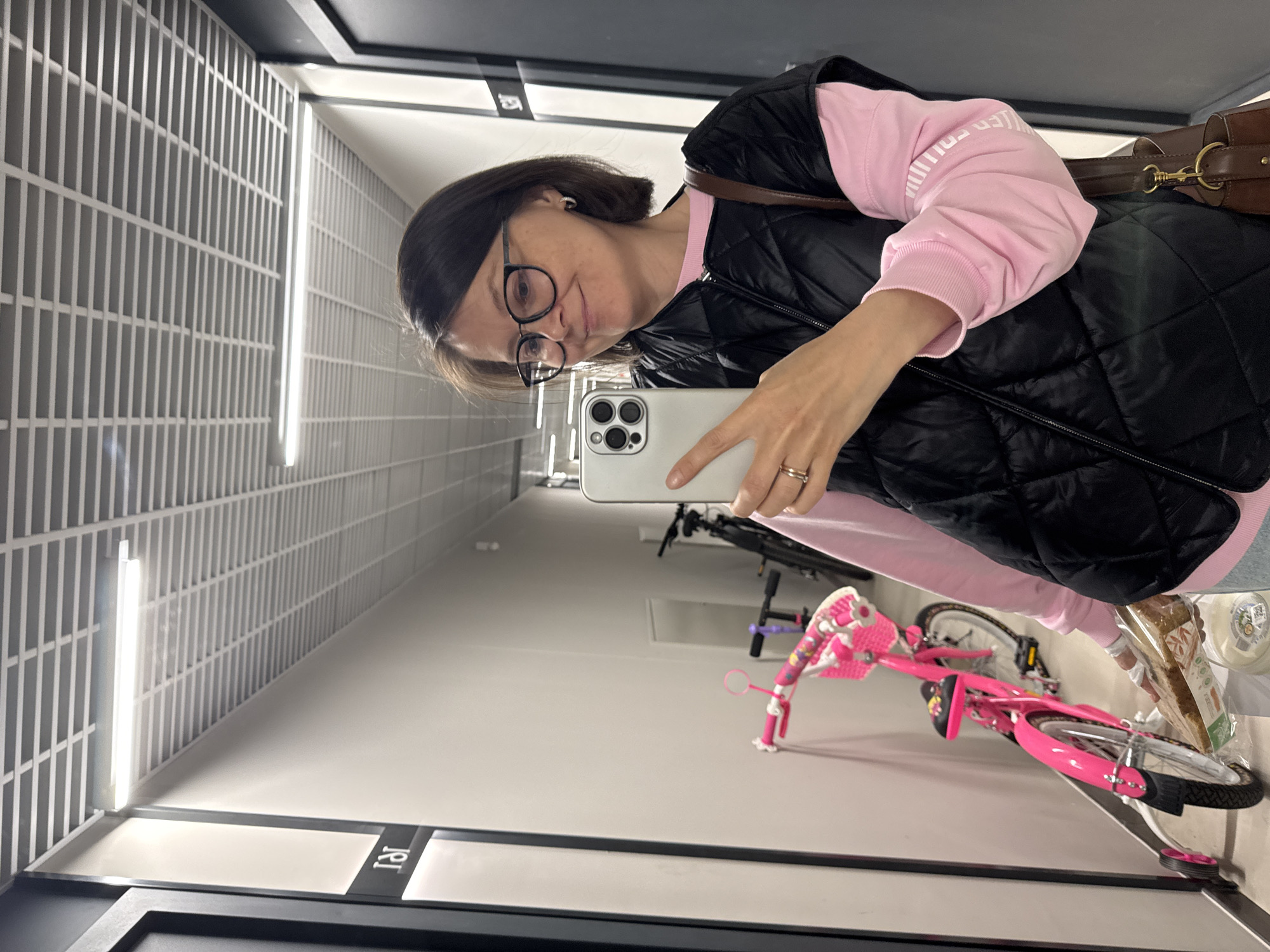 An upright, mirror selfie captures a woman posing in an indoor setting. The woman, appearing in the upper right quadrant, has dark hair, wears eyeglasses, a black puffer vest over a pink long-sleeved top, and a brown shoulder bag. Her left hand holds a white/silver smartphone with a triple camera setup, reflecting her image. A ring is visible on her ring finger. She has a slight, pleasant smile.
The setting appears to be a brightly lit, modern corridor or hallway, possibly in an apartment building or commercial space in Izhevsk, Russia. The walls are covered in light grey, rectangular tiles, and illuminated by vertical strip lights. The floor is light-colored and reflective.
In the lower left, reflected on the floor, the Russian word "ТЯГ" (TYAG), meaning "traction" or "pull," is visible on a dark strip.
Prominently featured in the lower half of the image are two bicycles. A bright pink child's bicycle with training wheels and a small basket on the handlebars dominates the lower right. Behind it, a dark-colored adult bicycle is partially visible. Near the rear wheel of the pink bicycle, there's a packaged item, possibly bread, and a white container.
On the woman's pink long-sleeved top, partially visible inverted text reads "SKILLER COLLIDE." The overall scene suggests a moment captured during an outing or within a residential building, with the bicycles indicating transportation or leisure.