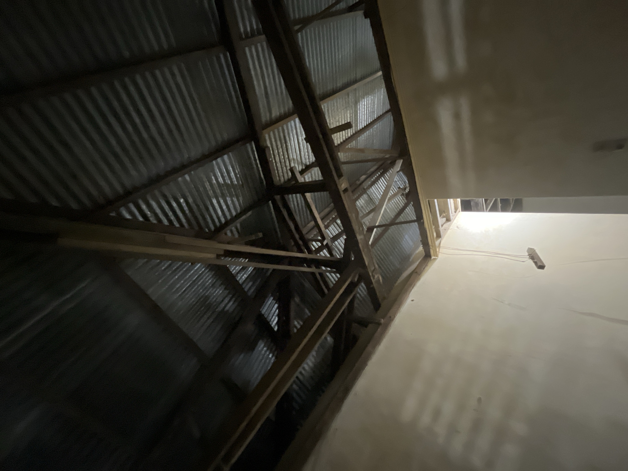 The image depicts the interior of an unfinished or damaged building, likely in the city of Kota Kuala Simpang, Indonesia. The focus is on the exposed roof structure, which consists of corrugated metal sheeting supported by wooden beams and rafters. The walls of the room are plain and appear to be plastered, with some areas showing discoloration or imperfections. A thin cord hangs from the ceiling, attached to a small wooden object, possibly part of an unfinished electrical installation or a hanging decoration. Light streams in from an opening, suggesting a window or an unfinished doorway, illuminating the dust motes in the air and casting shadows across the room and roof. The overall impression is one of an abandoned, under-construction, or post-disaster site, with the materials and construction style being consistent with the region.