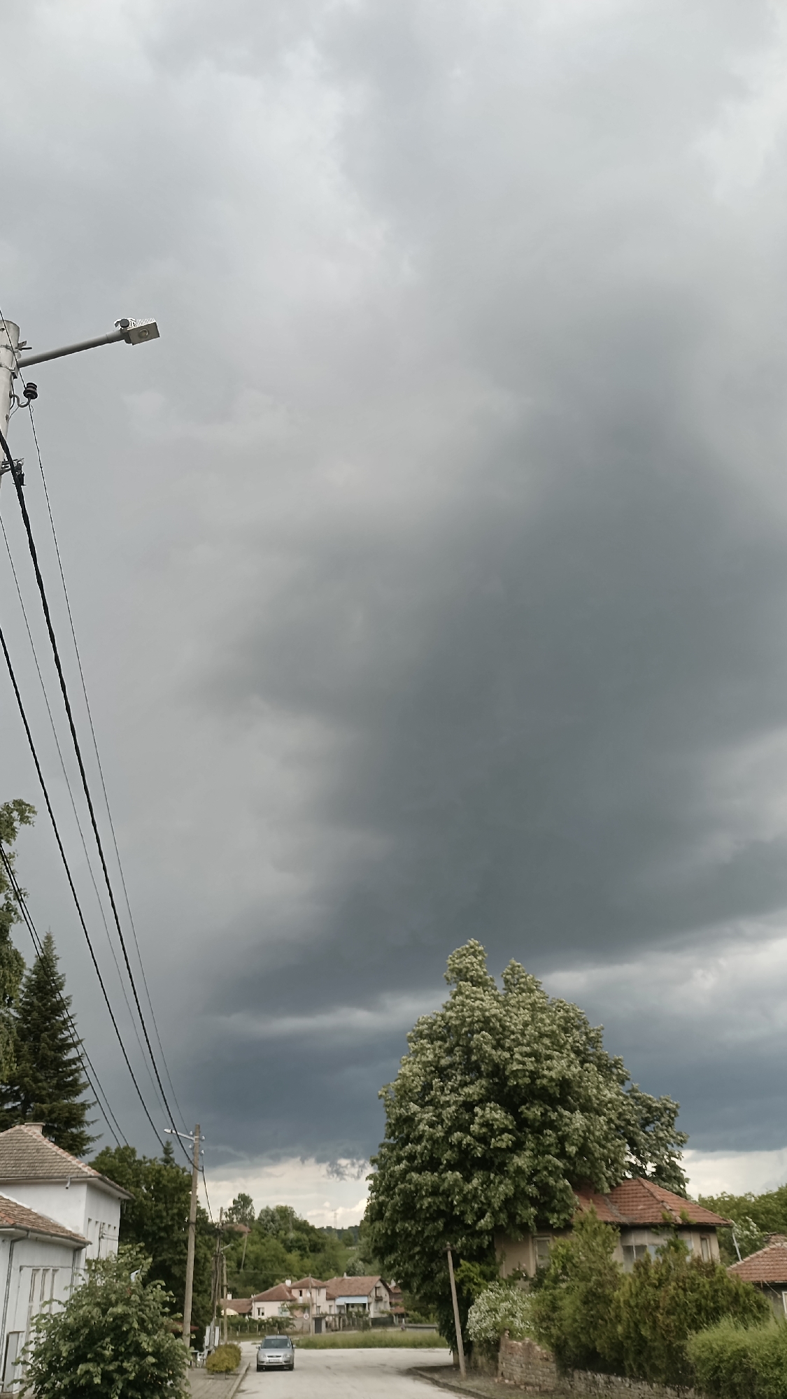 The image depicts a street in Muzga, Bulgaria, under a heavy, overcast sky. A silver car is positioned on the road. Houses line the street, partially obscured by trees. A utility pole with electrical wires is visible on the left side.