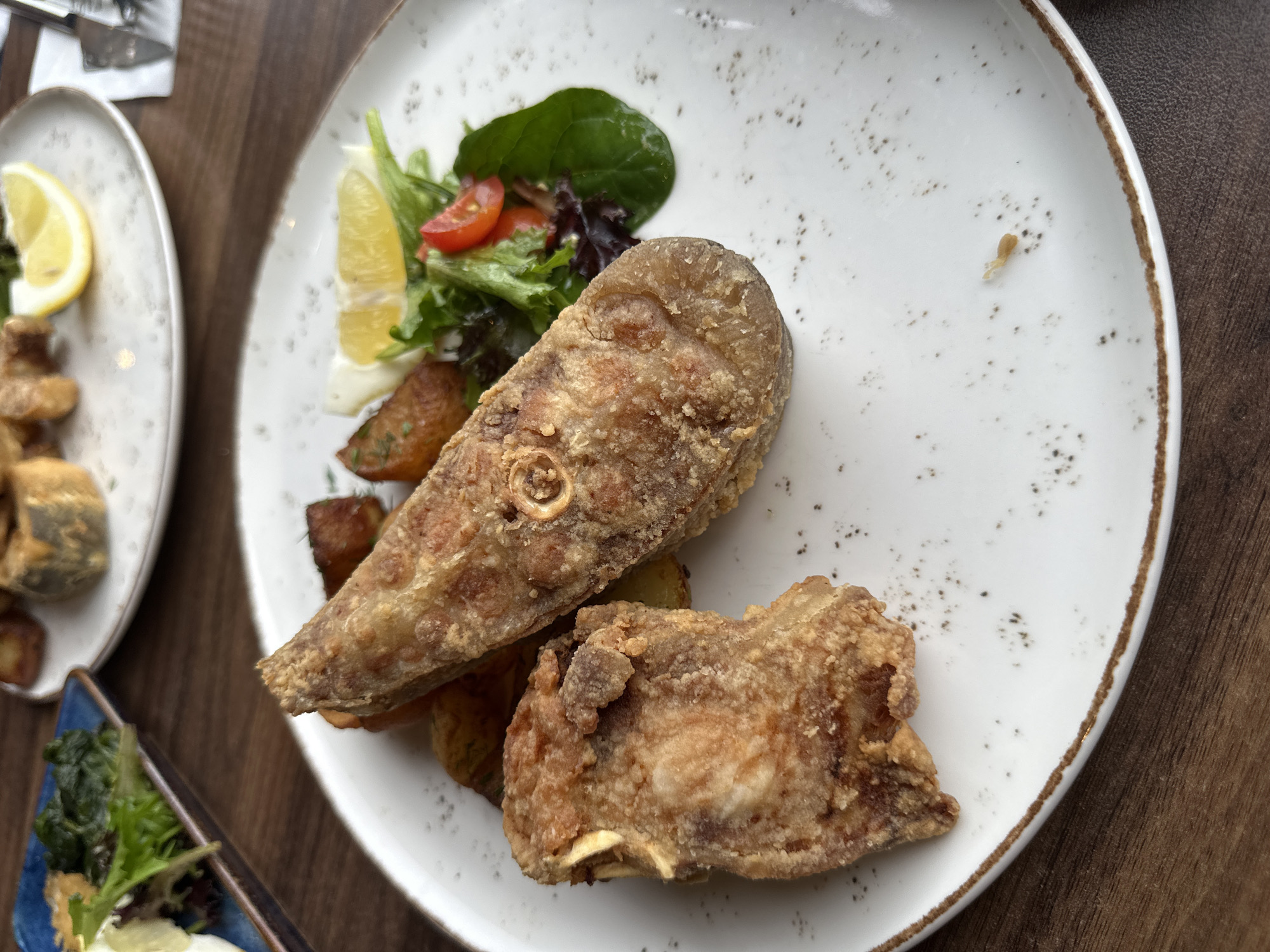 The image shows two white plates on a dark wooden surface. The plate on the right contains two pieces of fried fish served over potatoes, with a side salad containing lettuce, lemon, and tomatoes. The plate on the left contains fried pieces of similar food with lemon wedges, accompanied by side salad in a small blue container in the foreground. The image was likely taken indoors. The presence of Cyrillic characters cannot be confirmed, there is no evidence suggesting Vidin, Bulgaria as the location.