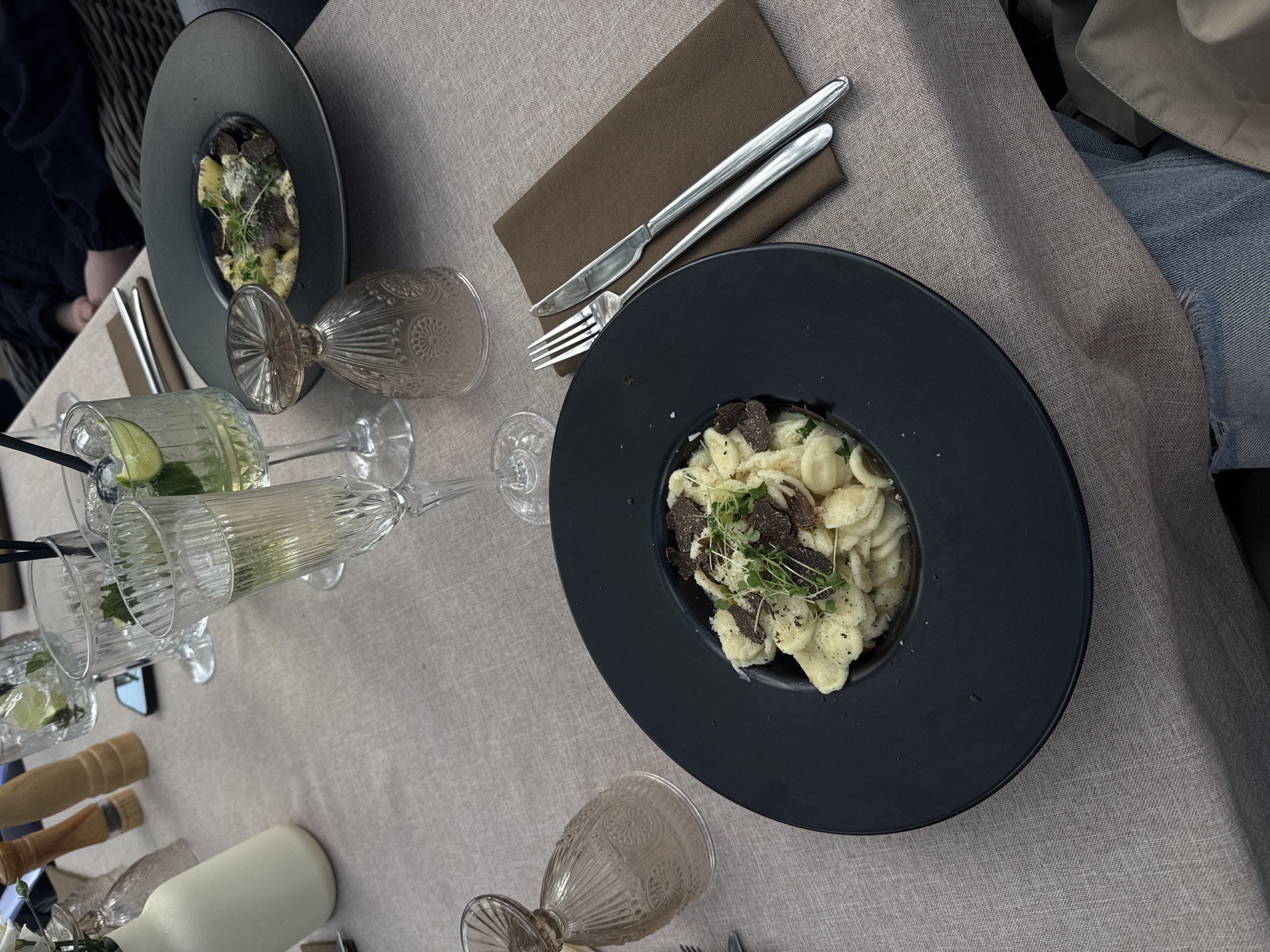 The scene depicts a dining table arrangement, likely in a restaurant, featuring pasta dishes topped with what appears to be truffle shavings. The location is Konstancin-Jeziorna, Poland, as indicated in metadata not provided but which can be deduced by the image set name.
On the table, there are two dark-colored plates, each containing a serving of pasta. Silverware (fork and knife) rests on a brown napkin beside one of the plates. Several glasses are visible, some filled with a clear liquid (possibly a cocktail) containing lime slices. Also visible are a bottle and other tableware items. The tabletop has a light, textured fabric covering. In the background, a person wearing jeans and a beige jacket is partially visible. Another person in a dark blue garment is seated on the left.