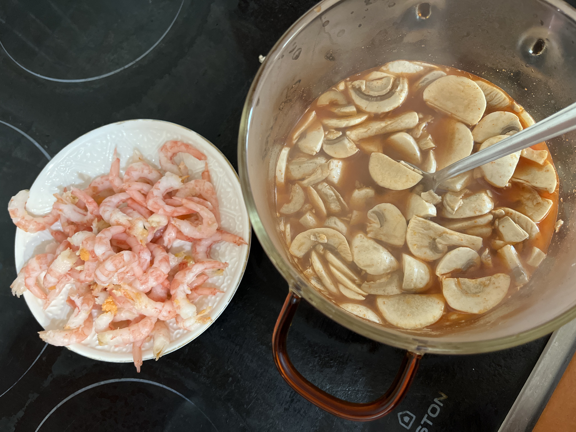 The image presents a culinary scene, likely indoors on a kitchen countertop, featuring ingredients for a dish. On the left is a white plate filled with peeled pink shrimp. To the right, there is a transparent glass pot containing a brownish liquid and a substantial quantity of sliced mushrooms. A metallic spoon rests within the pot, partially submerged in the liquid. The background indicates a black cooktop. There is no verifiable evidence to suggest that this image was taken in Chaikovsky, Russia.