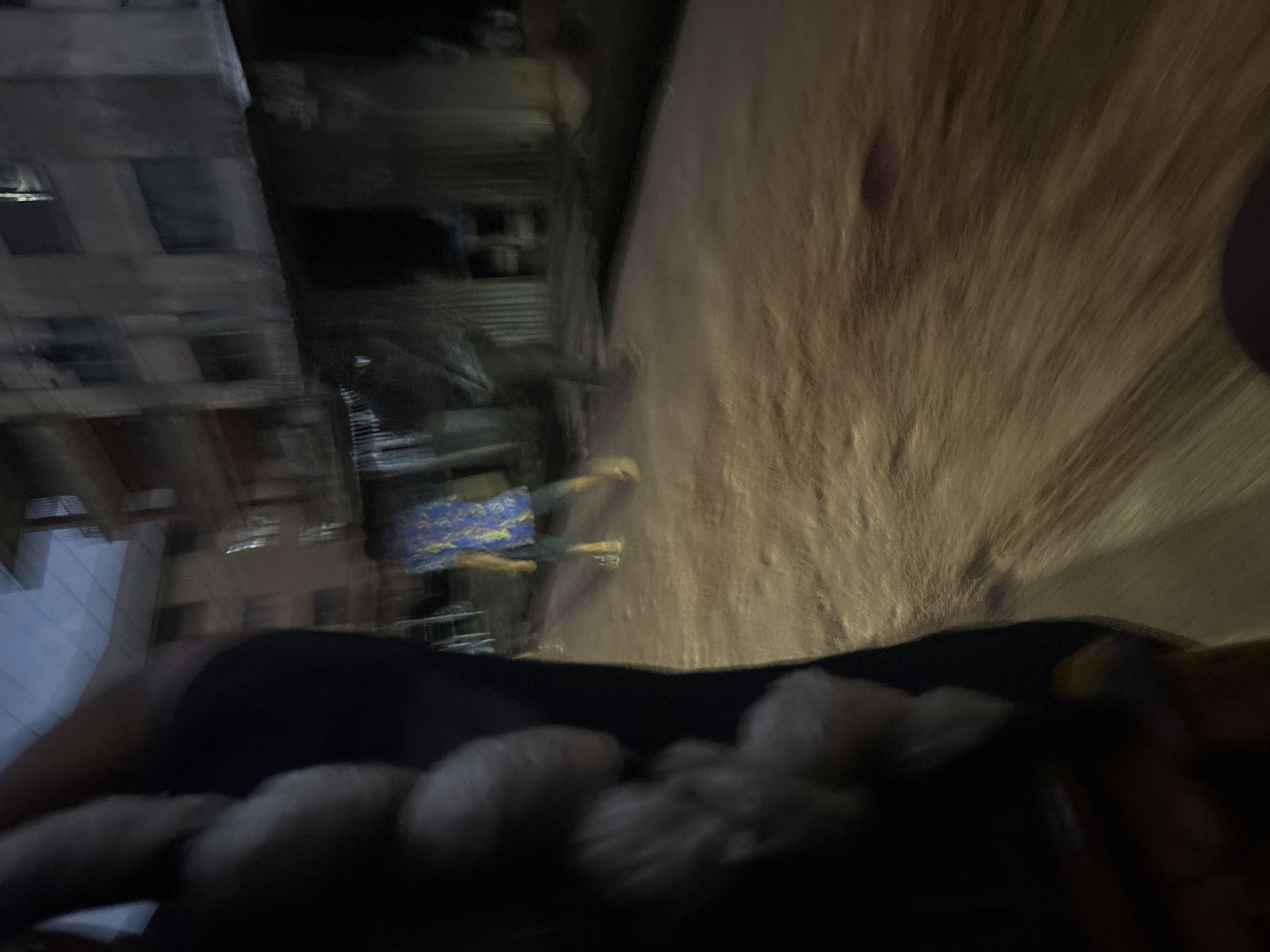 A nighttime scene in Onitsha, Nigeria, shows motion blur due to movement, suggesting a vehicle is in transit. In the foreground, a dark object, possibly a person's arm, is visible. Further back, a person in a blue patterned shirt and dark shorts walks on a dirt road. Buildings line the street, and the lighting suggests artificial illumination from streetlights or nearby structures. The overall impression is of a busy urban environment at night.