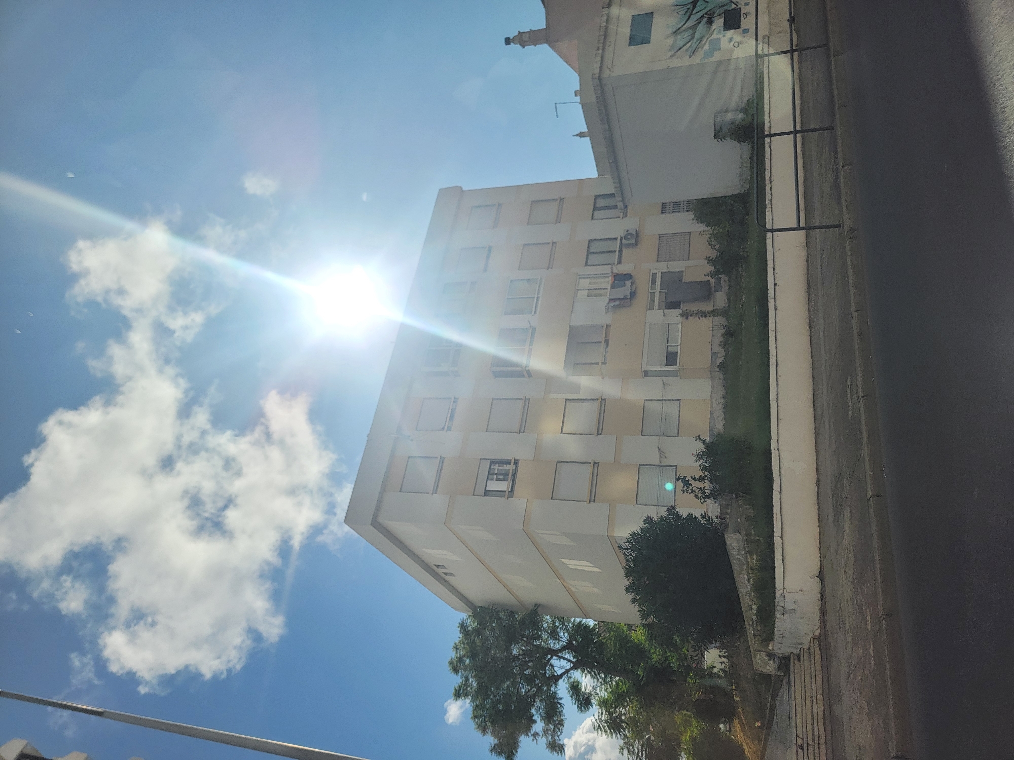 In Faro, Portugal, a five-story apartment building stands prominently under a bright, sunlit sky with scattered white clouds. The building, painted in pale yellow and cream, features numerous windows with closed white shutters, with a few exceptions displaying open blinds. Laundry hangs from a line outside one of the lower-story windows. Adjacent to the building on the left, a large, mature tree with dense foliage stretches upwards. A lower, darker green shrubbery borders the base of the apartment building, set behind a low, white-painted retaining wall. To the right of the apartment building, a smaller white building is partially visible, featuring a stylized mural on its side. In the foreground, a dark asphalt road runs horizontally across the bottom of the image, separated from a paved sidewalk by a curb. A black metal railing runs along the edge of the sidewalk. The sun's intense glare creates lens flare effects across the upper portion of the image. Stairs are visible leading up a grassy incline to the left of the apartment building.