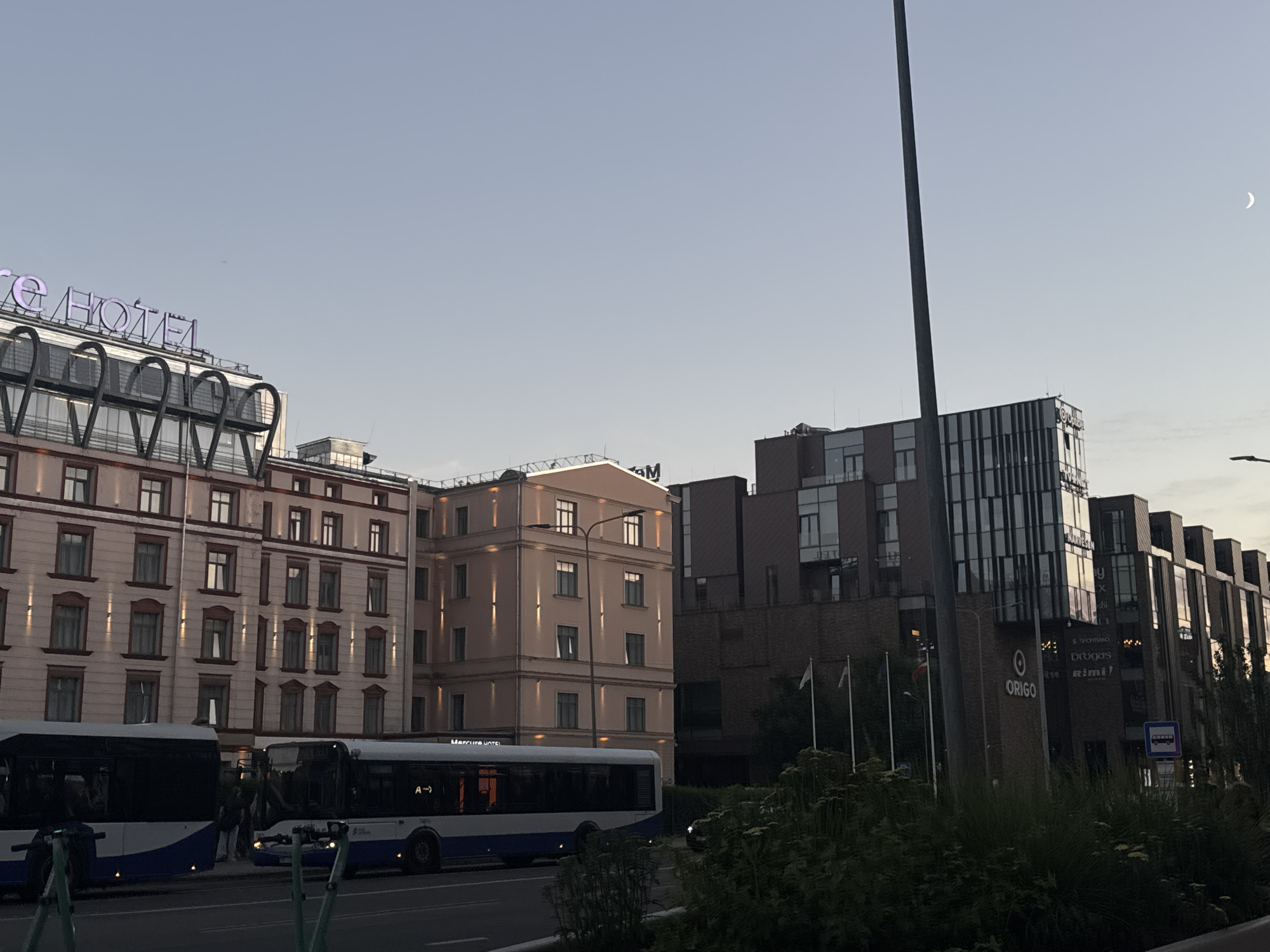 The image shows a street scene in Riga, Latvia, with several buildings and vehicles visible. In the foreground, two buses are parked on the side of the road. The bus on the left is dark and appears to be stationary, while the bus on the right is blue and white and also appears to be stationary. Behind the buses, a light pink building with multiple windows is visible, and to its right, a darker, modern building with a glass facade is present. A tall, dark pole stands prominently on the right side of the image, casting a shadow. A crescent moon is visible in the pale sky. The overall impression is that of a city street during daylight hours, possibly late afternoon or early evening. The scene is devoid of any explicit human activity or interaction.