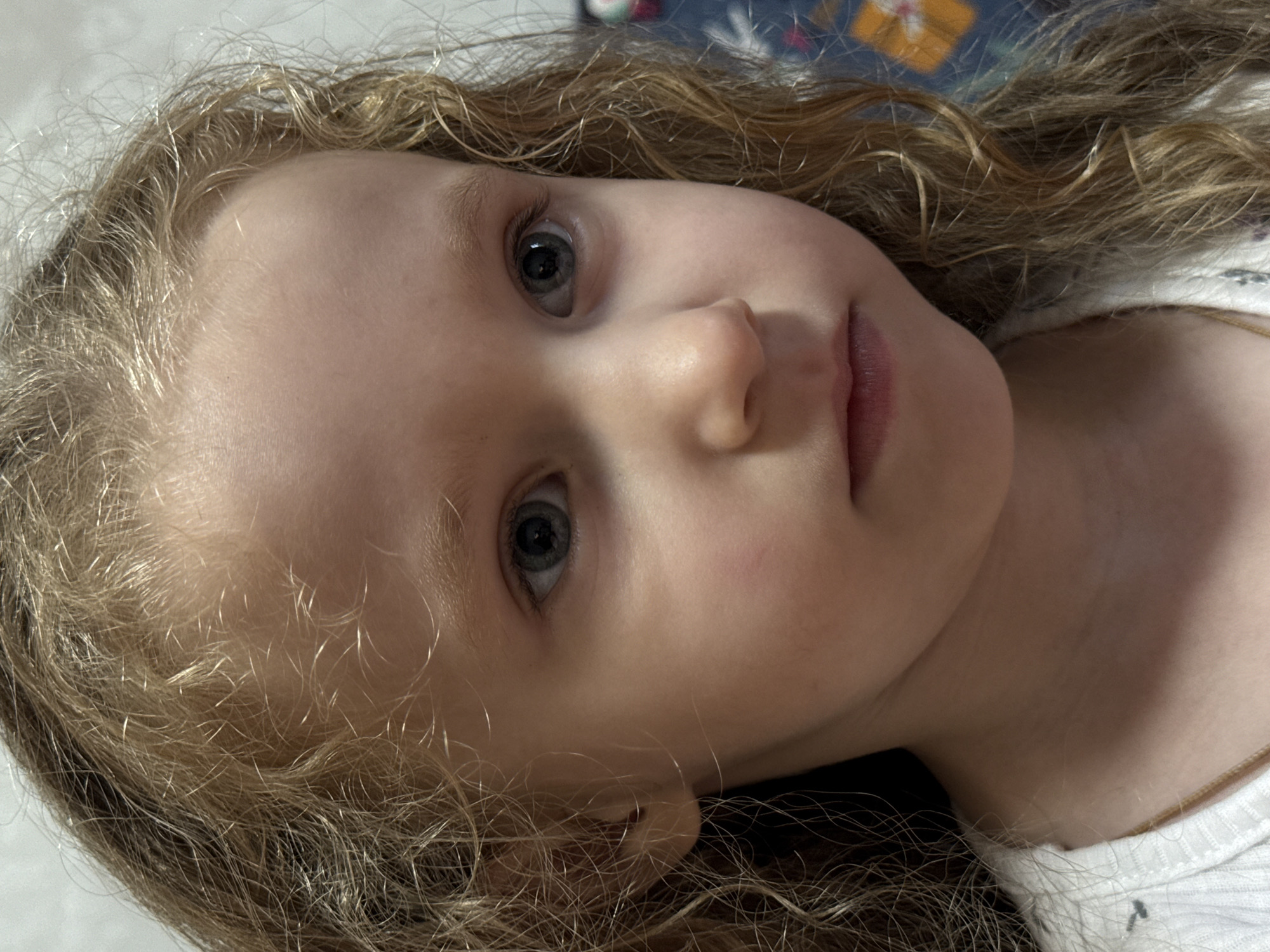 The image is a close-up portrait of a young girl with blonde, curly hair. She has light-colored skin, blue-gray eyes, and a neutral expression. She is wearing a white top. The background is blurred and appears to be indoors, possibly a room with a patterned fabric or wallpaper. There is no discernible information about the location within the city of Chaikovsky, Russia.