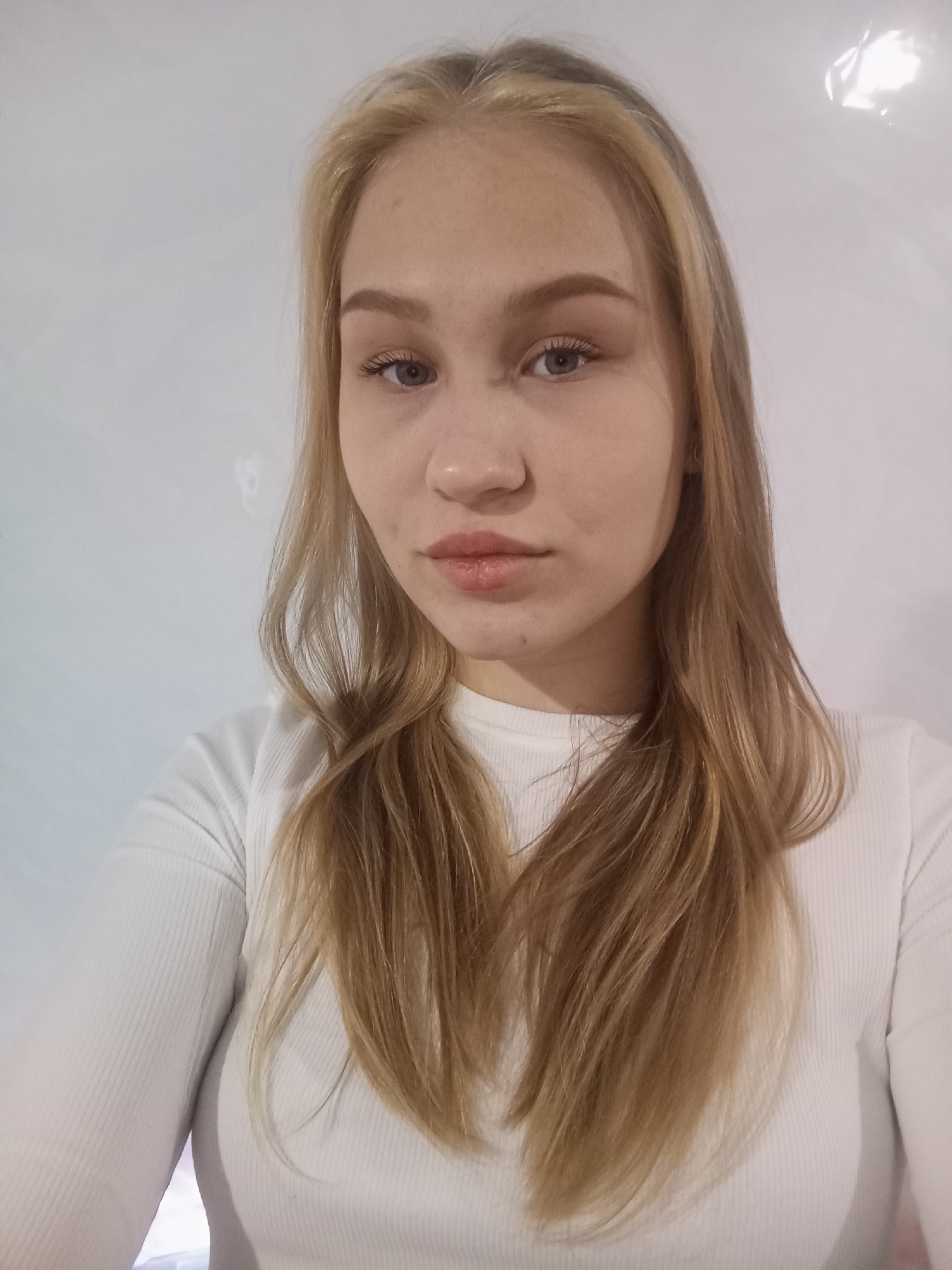 A close-up, front-facing portrait of a young woman with long, light-brown hair streaked with blonde highlights. She has fair skin, light eyes, and full lips. She is wearing a white, long-sleeved ribbed shirt. The background is a plain white wall. There is no indication of the city of Krasnodar, Russia, or any other location information visible in the image.