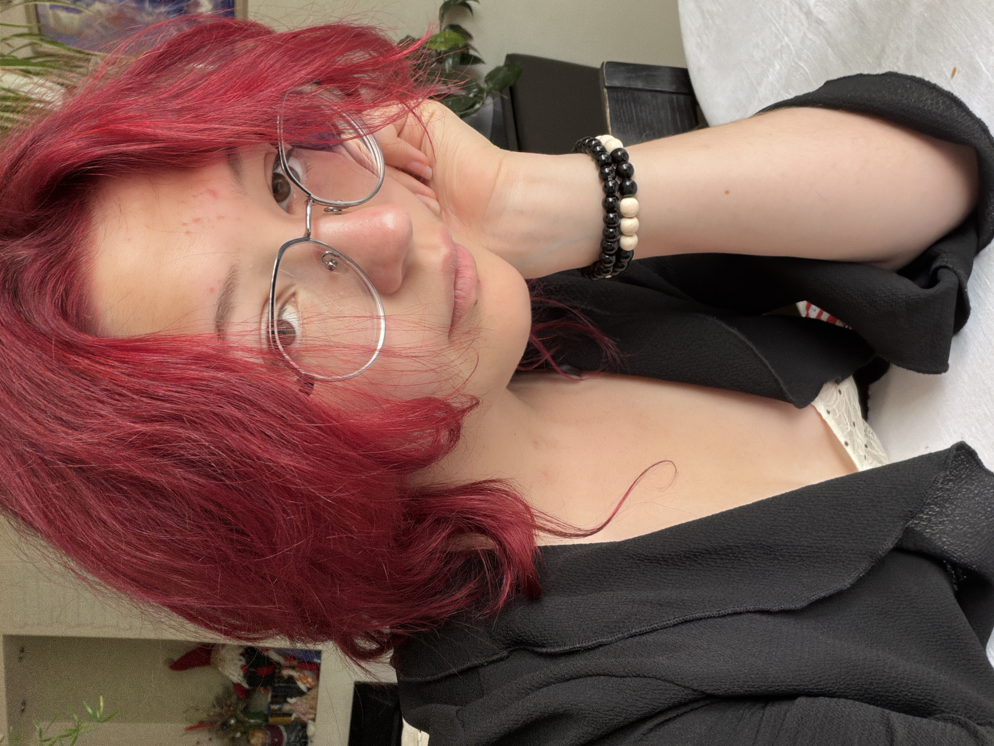 A young person with vibrant red hair, wearing silver-rimmed glasses and a black blazer over a white lace top, rests their head on their hand. They are adorned with two bracelets on their wrist: one with black and light-colored beads, and another entirely of black beads. The scene takes place indoors, likely in an apartment or home setting, with a white tablecloth covering a surface in the foreground. In the background, shelves hold various decorative items, including figurines and what appears to be a plant. The overall lighting suggests daytime. There is no visible indication of the city of Chelyabinsk, Russia.