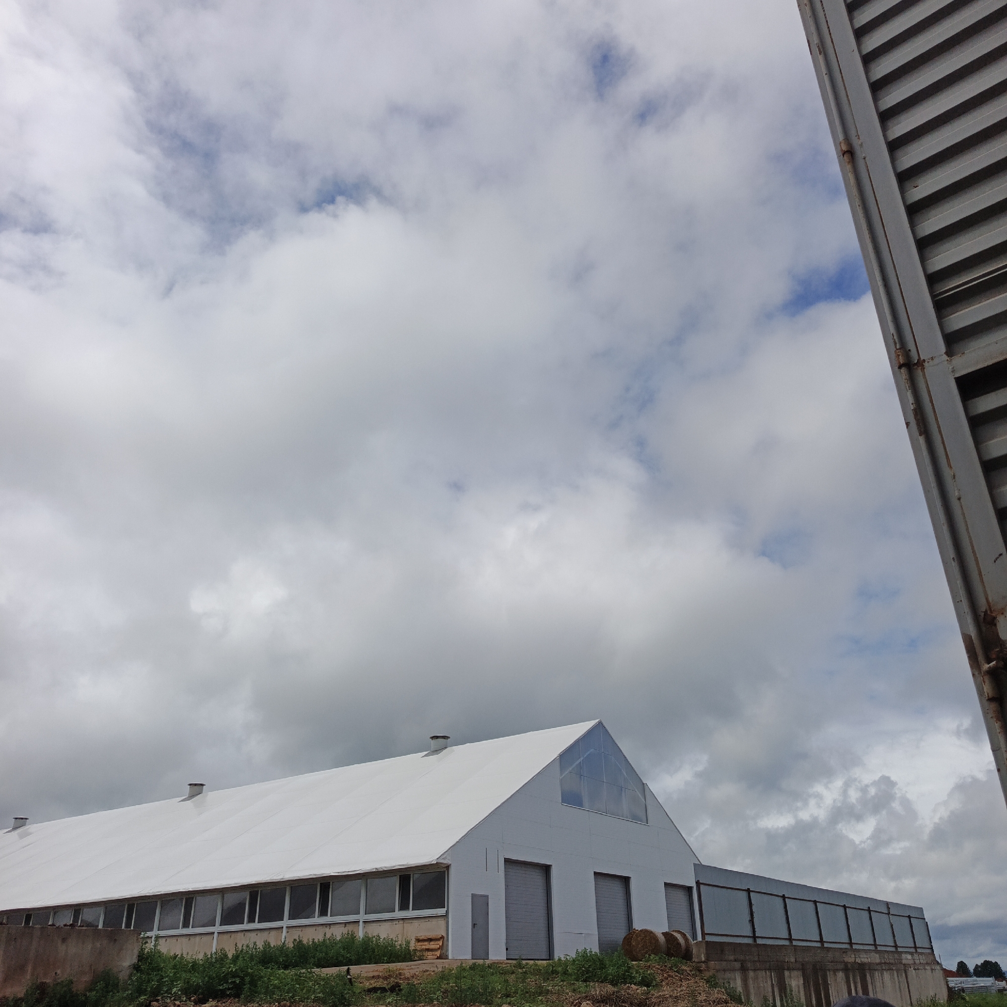 The image depicts a large, white, tent-like structure, likely a barn or agricultural building, under a cloudy sky. The building has a long, sloped roof and a front facade with several large garage doors. Large windows run along the side of the building, and a triangular section at the front appears to be made of a translucent material. Two circular hay bales are visible near the front of the building, and green vegetation grows around the base. To the right of the frame, a portion of a corrugated metal structure, possibly a shipping container or part of another building, is visible. The sky is filled with dramatic, grey and white clouds, suggesting overcast or possibly stormy weather. The overall scene conveys a sense of rural or agricultural setting, possibly under construction or in use for farming. No people are visible in the image. There is no visible text.