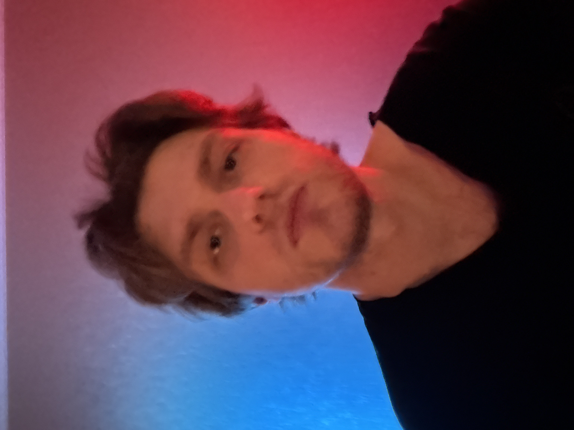 This is a close-up, low-angle, vertically oriented, indoor photograph of a young adult male. He is looking directly at the camera with a neutral expression. The man has light skin, brown hair, and a light beard. He is wearing a black t-shirt. The background is softly lit with a gradient of red on the top and blue on the bottom, suggesting artificial lighting such as colored LED lights. The lighting creates a slightly diffused and atmospheric effect on the subject's face and hair. There is no visible text in the image. The overall impression is of a casual, self-taken photograph.