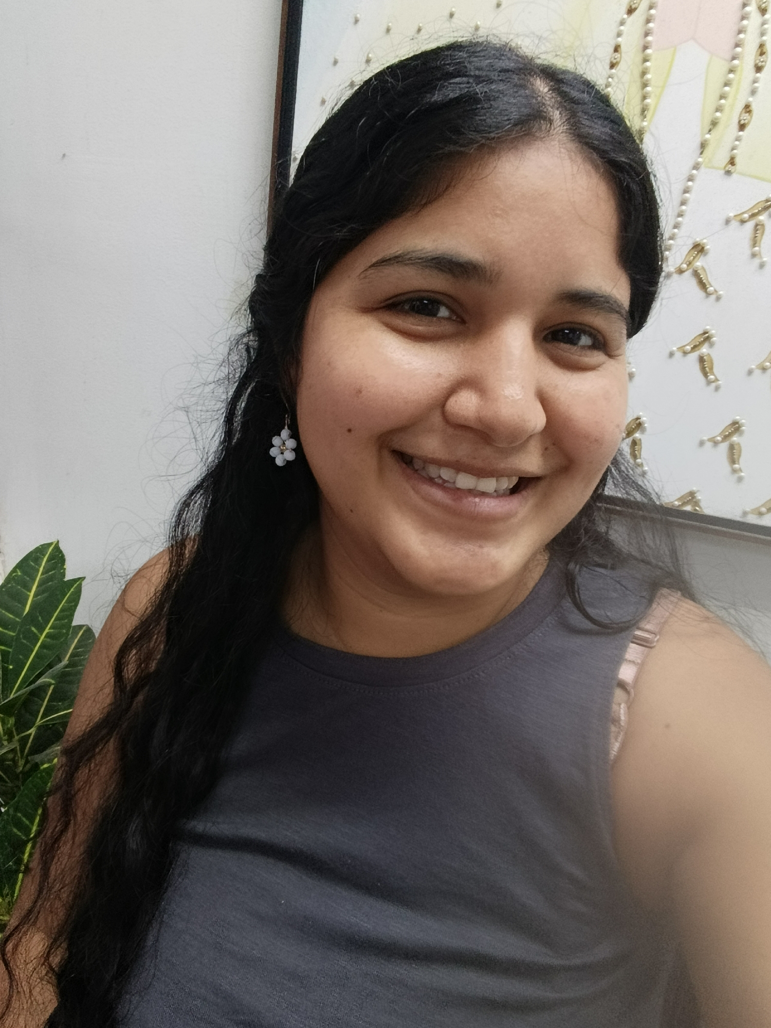 A young woman with long, dark, wavy hair is looking at the camera and smiling. She is wearing a dark gray tank top and a delicate, light blue flower-shaped earring on her left ear. The background is a white wall with some artwork or decoration on it, featuring pearl-like embellishments. To the left of the woman, a portion of a plant with green leaves is visible. The lighting suggests an indoor setting. The image is a selfie, taken from a slightly high angle.