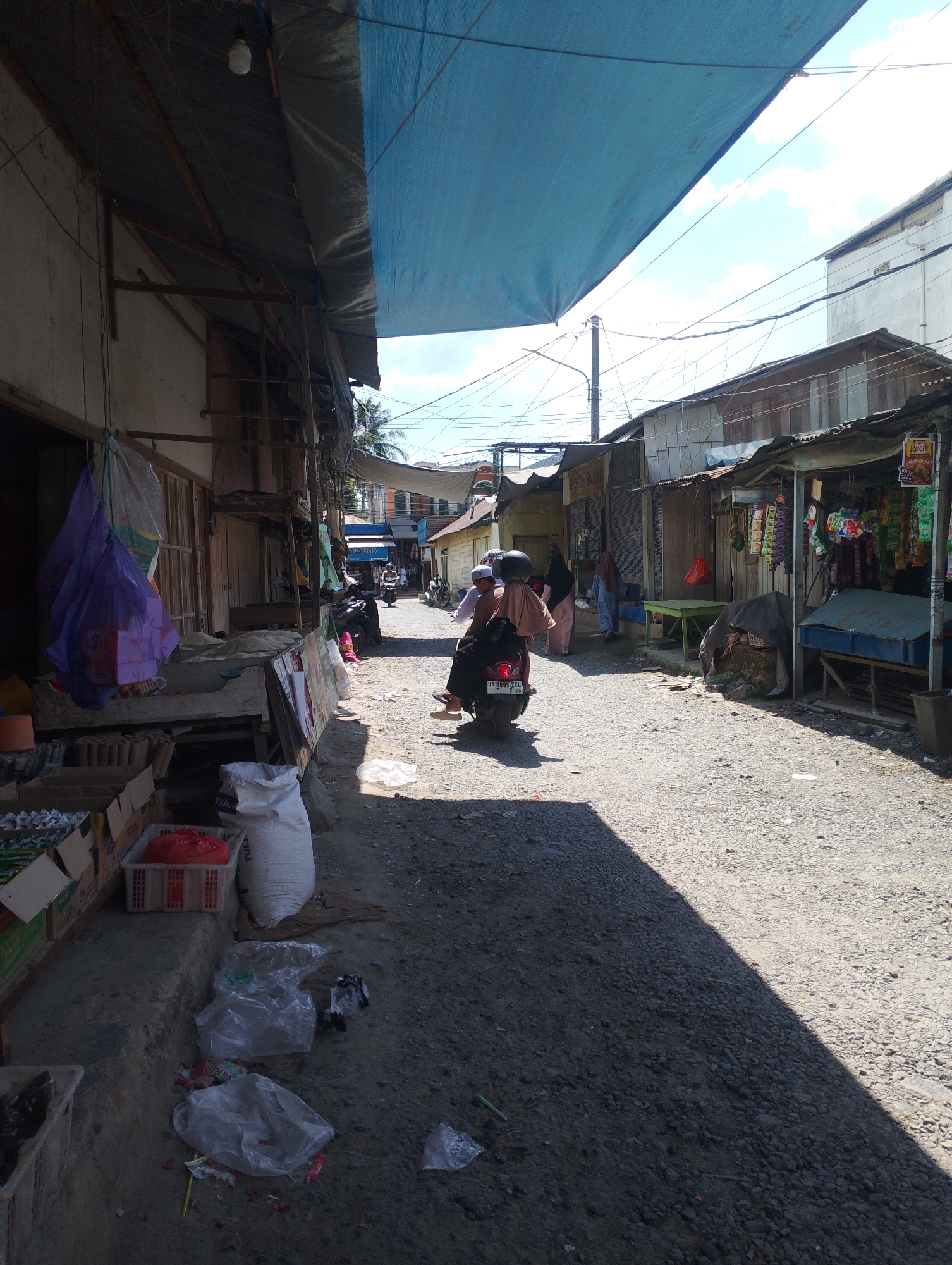 The image depicts a street scene in Tungkaran Pangeran, Indonesia, likely a market area. A person is riding a red motorcycle with a passenger, both wearing dark clothing, down a gravel road. The road is lined with open-air stalls or small shops with corrugated metal roofs and awnings, some covered with blue tarpaulin. The stalls display various goods, including packaged items hanging from racks and items in boxes and baskets. Several people are visible walking in the background. Trash, including plastic bags and wrappers, is scattered on the ground and sidewalk. The weather appears sunny, casting strong shadows across the road.