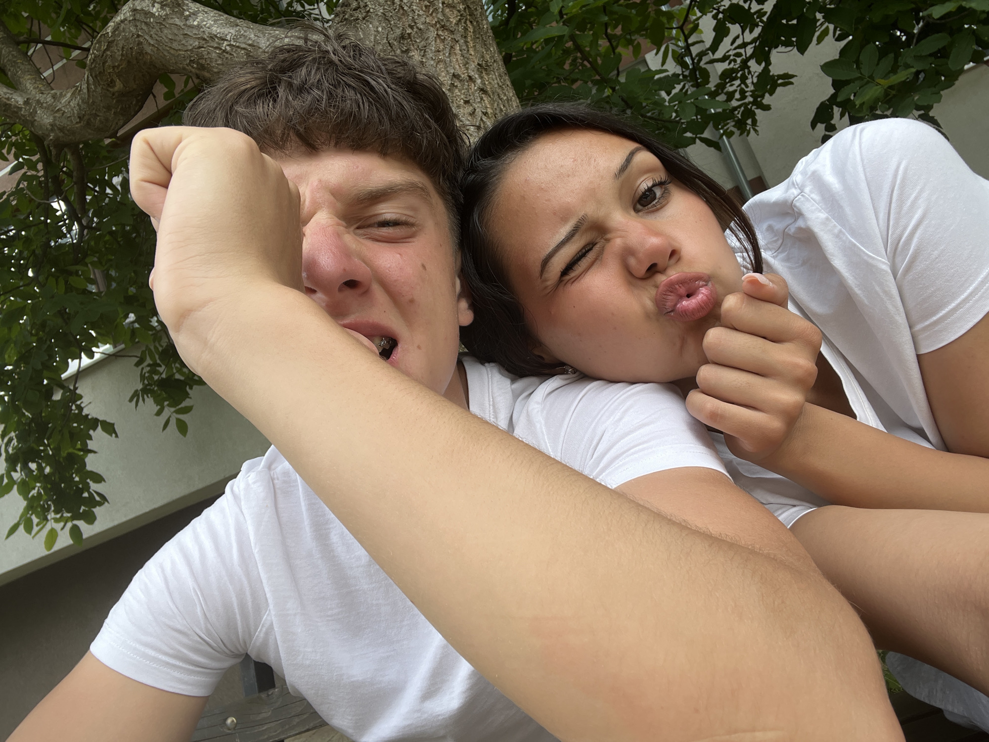 This is a close-up, selfie-style photograph taken outdoors, likely during the daytime. The scene features two young people, a male and a female, with a tree and foliage in the background. The female subject is positioned behind and to the right of the male subject. Both are wearing white t-shirts.
The male subject is in the foreground, his face close to the camera. He has dark, messy hair and is making a playful grimace, with his right fist held up near his face, obscuring part of his eye. His mouth is open, revealing braces on his teeth. The female subject is leaning on his shoulder or arm. She has dark hair and is winking with her left eye, while puckering her lips in a kissy-face pose. Her right hand is near her face, with fingers extended.
The background suggests an outdoor setting, possibly a garden or park, with the textured bark of a tree and green leaves visible. The lighting appears natural, consistent with daylight. The overall mood is lighthearted and playful, capturing a moment of shared amusement between the two individuals. There is no visible text in the image. The location context provided is Zlatograd, Bulgaria, which is consistent with an outdoor setting.