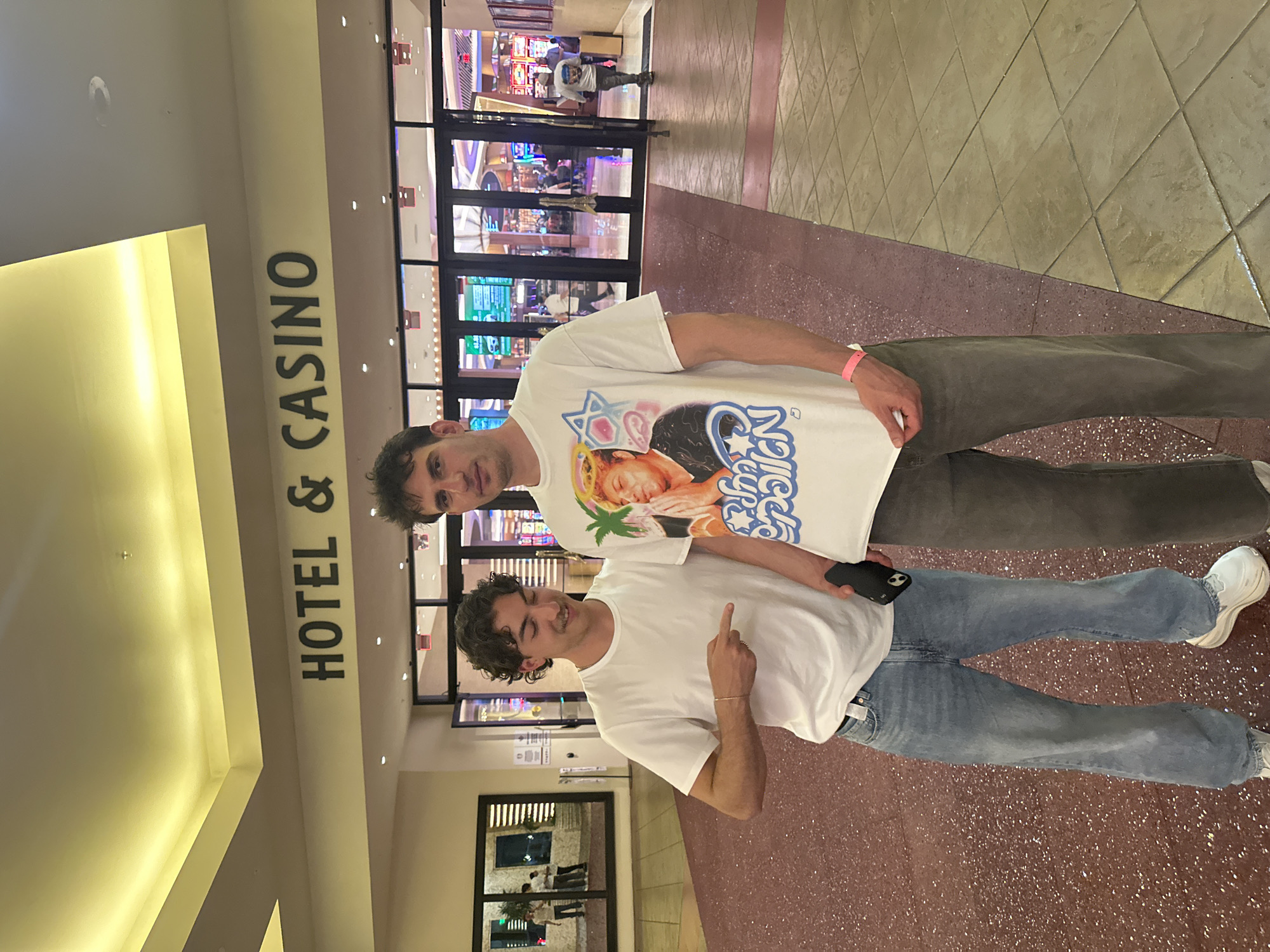 Two men are standing in front of the entrance to the Seminole Hard Rock Hotel & Casino in Hollywood, Florida. One man is wearing a white t-shirt with an image on it and grey pants. The other man is wearing a plain white t-shirt and jeans. The entrance to the casino is visible in the background.