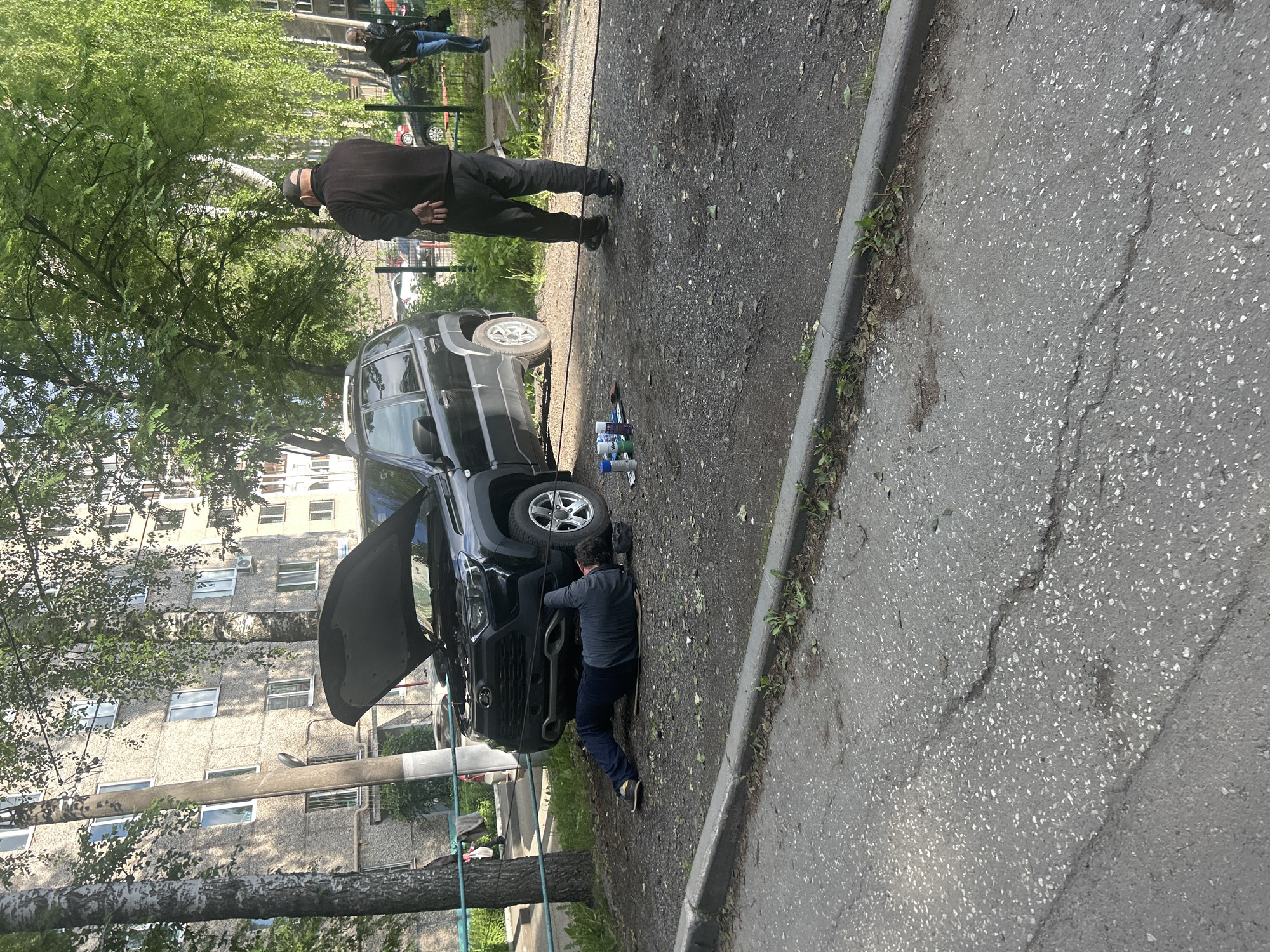 A daytime outdoor scene in Izhevsk, Russia, depicts two men engaged in vehicle maintenance or repair beside a multi-story apartment building. The image shows a dark-colored SUV or pickup truck parked on an asphalt surface with its hood open.
One man, wearing a grey long-sleeved shirt and dark pants, is kneeling on the ground in front of the vehicle, actively working on its front end or engine compartment. Several spray cans or bottles, possibly containing automotive fluids or cleaners, are placed on the ground near the front passenger wheel. A dark hose or cable runs along the ground towards the vehicle.
Standing upright behind the working man, closer to the rear of the truck, is another man dressed in a dark jacket, dark pants, and a dark cap. He has his hands clasped behind his back, appearing to observe the activity or wait.
The setting includes an asphalt ground, which is uneven and cracked in places, and a concrete curb along the right side of the frame. To the left, several trees with dense green foliage suggest a spring or summer day. In the background, a large, concrete apartment building with numerous windows, characteristic of Eastern European architecture, rises. Further in the distance, two other figures are visible. The lighting is bright, indicating clear or partly cloudy weather. No discernible text is visible in the image.