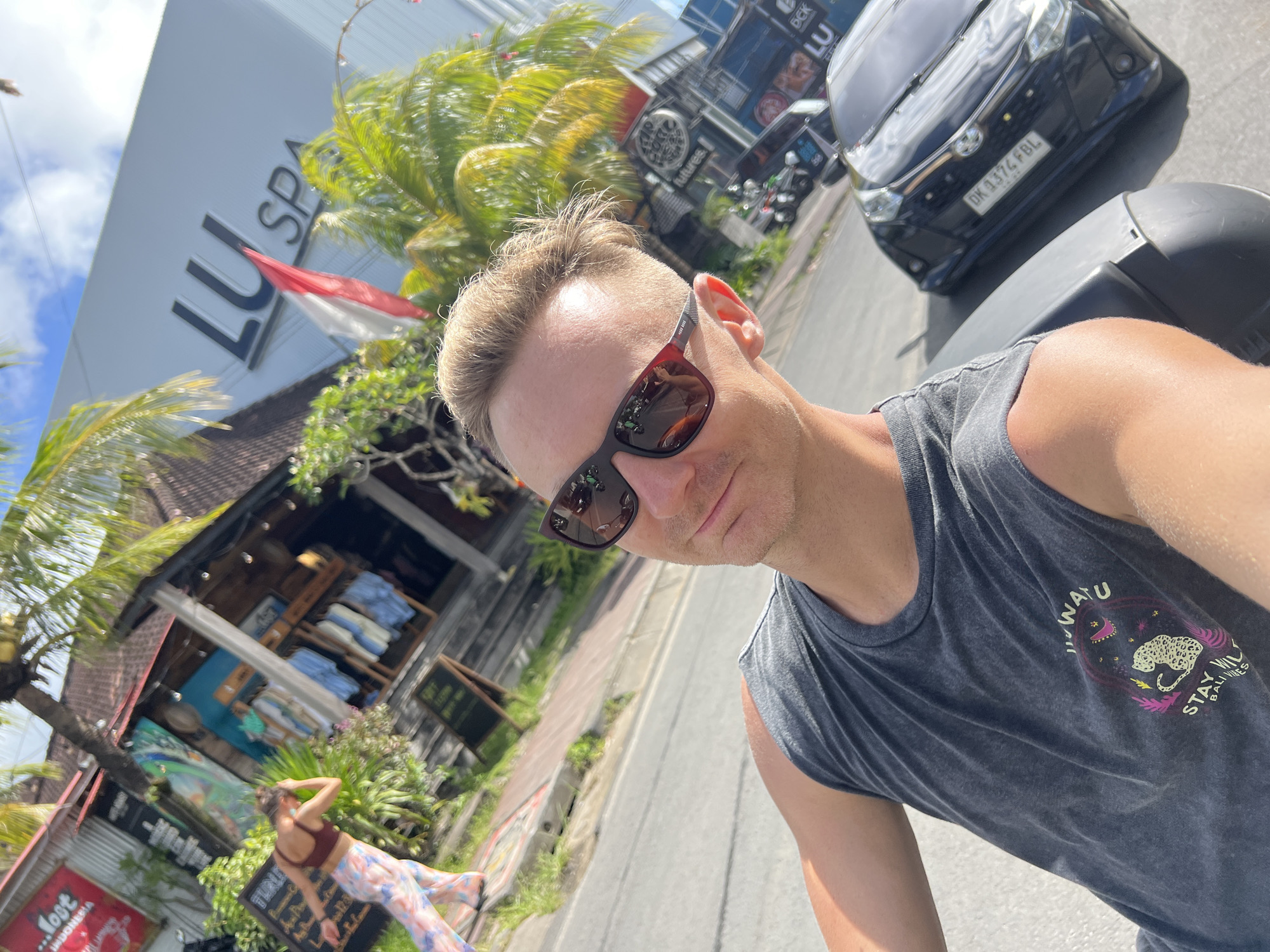 A man in sunglasses and a tank top is taking a selfie while on a motorbike in Kuta Selatan, Indonesia. The license plate DK 1374 FBL is visible on a black car to the right. Behind the man is a building with "LU SPA" on it and a waving Indonesian flag. To the left, a woman is standing in front of a business. Signage visible includes the words "Most" and "INDONESIA", "futures", "WATU", "STAY WILD", "BALLIKS", "LU", "BEK", and "TURLI".