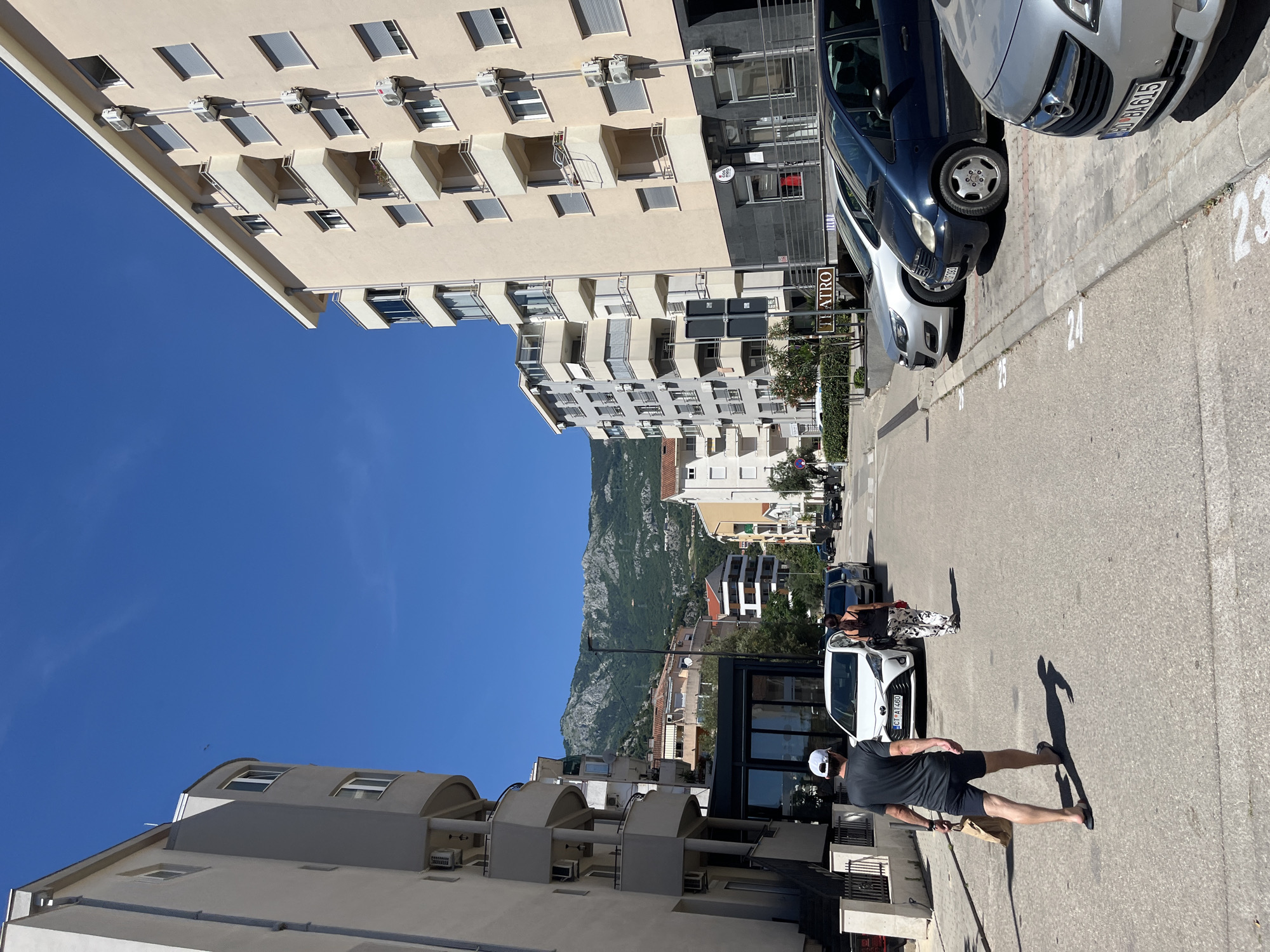 In Budva, Montenegro, on a sunny day, a street scene unfolds with residential buildings lining both sides of a paved road. The buildings are modern, characterized by light-colored facades and balconies. On the right side of the street, several parked cars, including a silver Opel, a dark blue Mercedes-Benz A-Class, and a silver sedan, occupy designated parking spots marked with white numbers. The backdrop features green, treed mountains under a clear blue sky. In the foreground, a man wearing a dark t-shirt, black shorts, a white cap, and flip-flops walks away from the camera, carrying a paper bag. He is followed by a woman wearing a dark top and patterned pants, walking in the same direction. The environment appears to be a residential or mixed-use urban area.