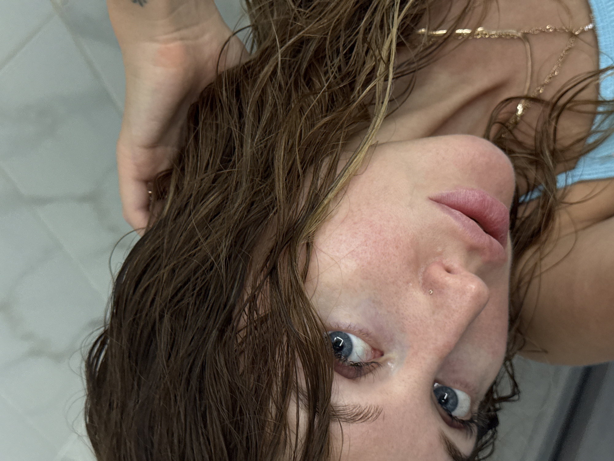 A close-up, inverted portrait features a young woman with wet, brown hair, looking directly into the camera. Her face is positioned upside down, with her blue eyes gazing upwards. Her light-toned skin shows natural freckles and a slight flush, and a delicate nose stud is visible on her right nostril. Her pink lips are slightly parted.
Her wet, medium to dark brown hair, which appears shoulder-length or longer, cascades around her face and shoulders, suggesting she might have recently showered or been in water. One hand, holding the back of her head, is partially visible in the upper left of the frame, revealing a small, dark tattoo on the wrist.
A thin, golden, beaded chain necklace or body jewelry is draped over her right shoulder and neck area. A light blue garment, possibly a top or swimsuit, is visible on her upper body. The background consists of light-colored, tiled surfaces, hinting at an indoor setting such as a bathroom or shower. Despite the unusual perspective, the woman's gaze is direct and engaging. There is no visible text in the image. The scene is situated in Chaikovsky, Russia.