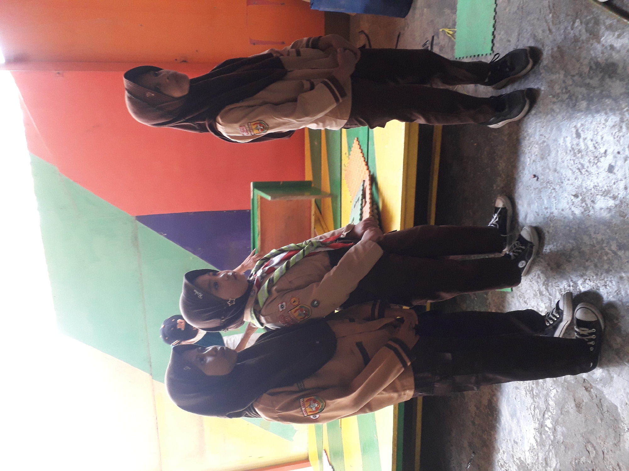 In a brightly lit indoor setting, possibly a school or community center in Cankuang, Indonesia, three young girls stand facing to the right, dressed in scout uniforms. The girl on the left wears a brown uniform with a dark headscarf and black sneakers. The middle girl, slightly further back, also wears a brown uniform with a dark headscarf and a scout scarf around her neck, paired with black sneakers. The girl on the right, partially obscured by the middle girl, is dressed in a similar brown uniform with a dark headscarf and black sneakers. They are positioned on a raised platform with a yellow base and a green and orange backdrop featuring geometric shapes. The floor is concrete, with scattered green mats visible. One of the girls has a badge on her uniform that appears to read "PRAMUKA RAJAWALI". The scene is static, suggesting a moment captured during an event or activity.