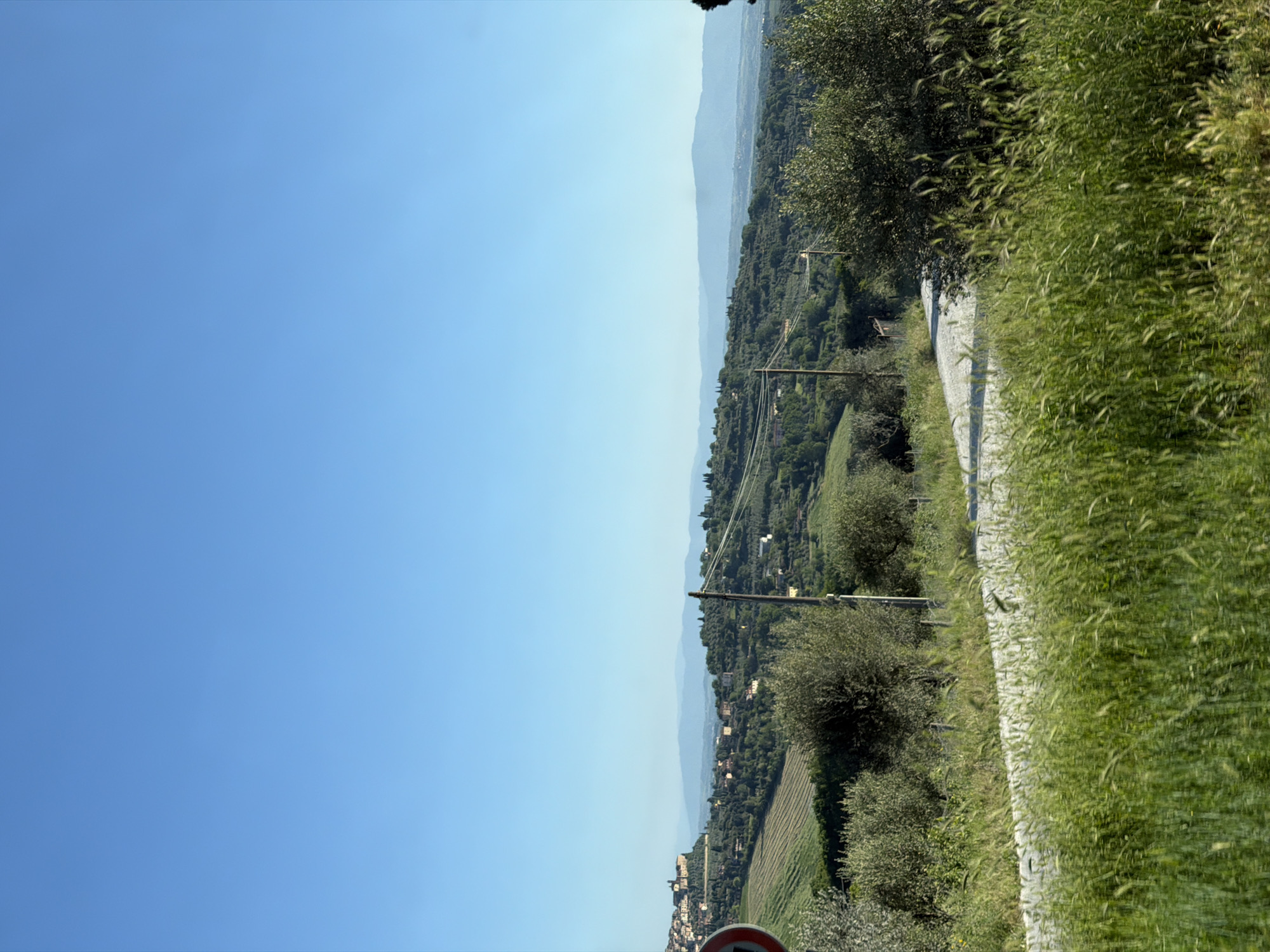 The image depicts a landscape in Chianciano Terme, Italy. The sky is clear and blue. A panorama of hills and mountains extends to the horizon. A road with overgrown grass on the sides runs along the right side of the frame. Utility poles are visible amidst the trees and vegetation. There is a red and white sign at the bottom of the image. Buildings are visible on the hillsides.