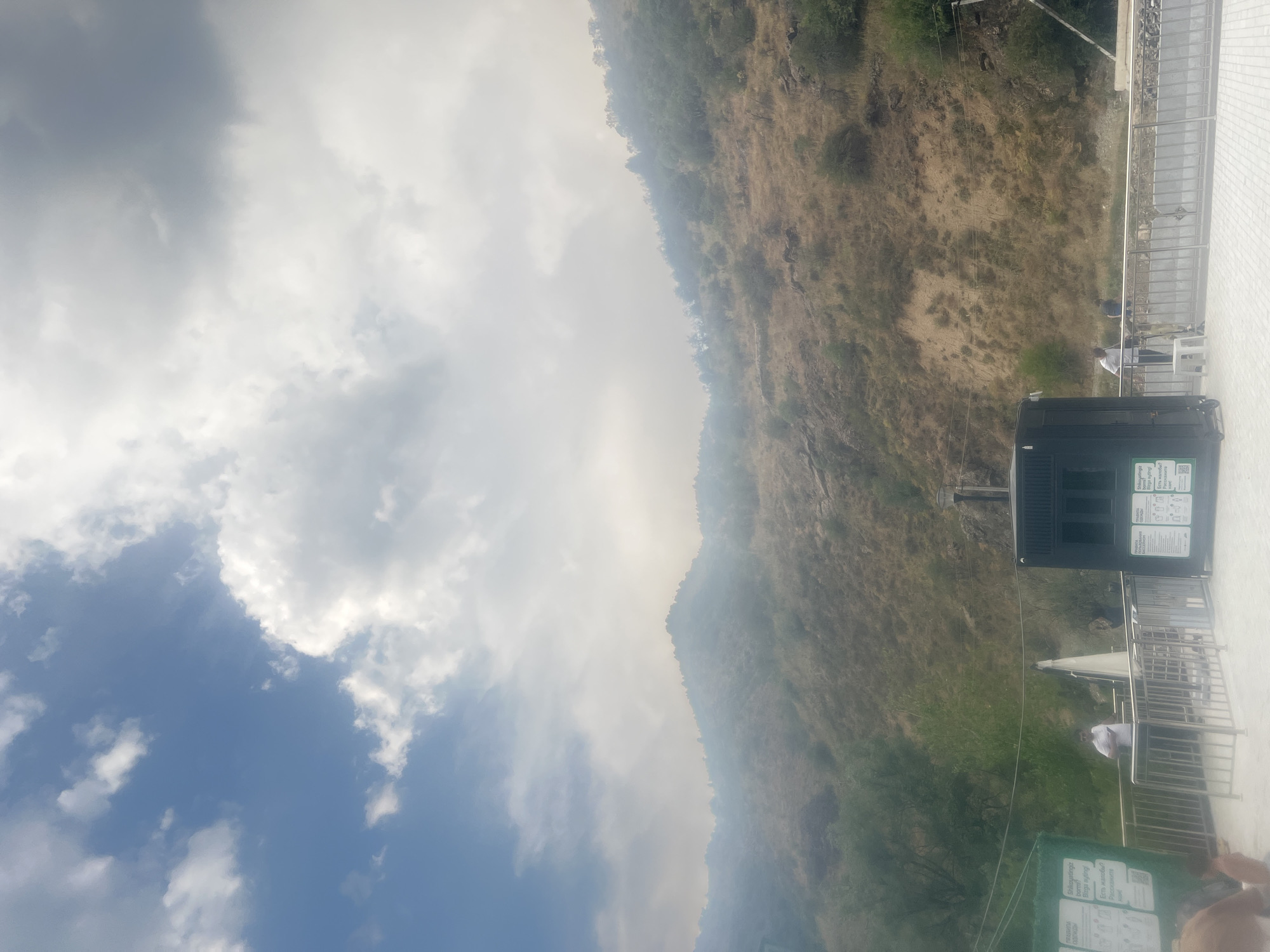 A wide-angle outdoor daytime shot captures a mountainous landscape under a partly cloudy sky. In the foreground, a paved area with metal railings separates people from the edge of the area. To the left, a green sign with white text is visible. A dark, rectangular structure with windows stands in the middle ground, accompanied by a white conical object behind it. A few individuals are present in the scene, some standing near the railings and others in the distance. The terrain behind the railing is a dry, grassy hillside dotted with sparse vegetation and some scattered trees. The overall scene suggests a public area or viewpoint in a natural, hilly environment.