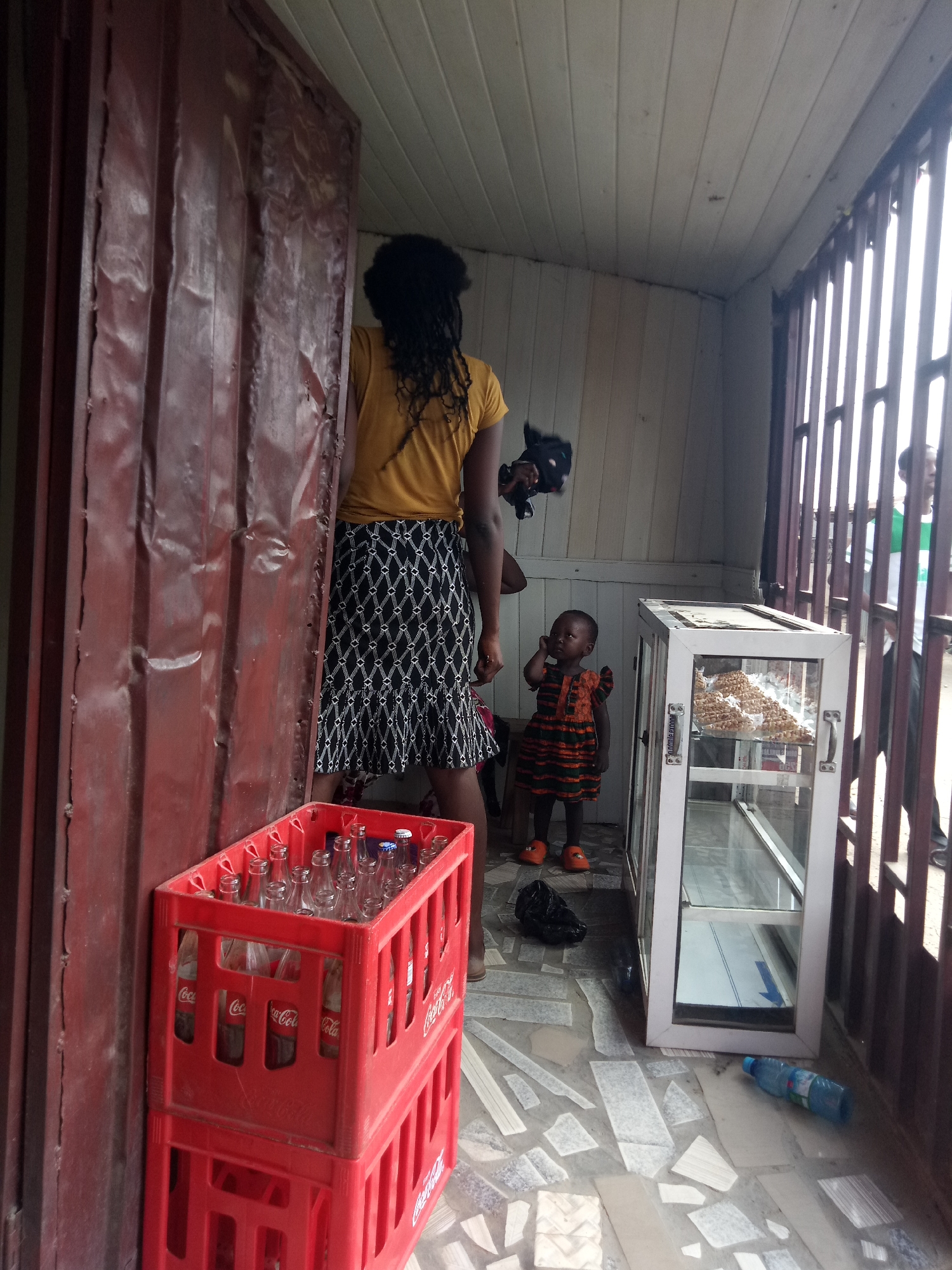 The image depicts an outdoor stall or kiosk with a corrugated metal structure on the left and a barred opening on the right. In the foreground, stacked red plastic crates filled with empty Coca-Cola bottles are visible. A woman with dark, braided hair, wearing a yellow t-shirt and a black and white patterned skirt, stands with her back to the viewer, facing into the stall. A young child in an orange and green striped dress and orange shoes stands near a glass display case. The display case is white and contains what appears to be a pile of nuts or snacks on the top shelf. The floor is tiled with a mosaic pattern of light-colored stones. A black plastic bag lies on the floor near the child, and a blue plastic water bottle is on the floor to the right of the display case. In the background, through the bars of the stall, another person wearing a green and white shirt is partially visible. The overall scene suggests a small business or market stall in an urban environment.
