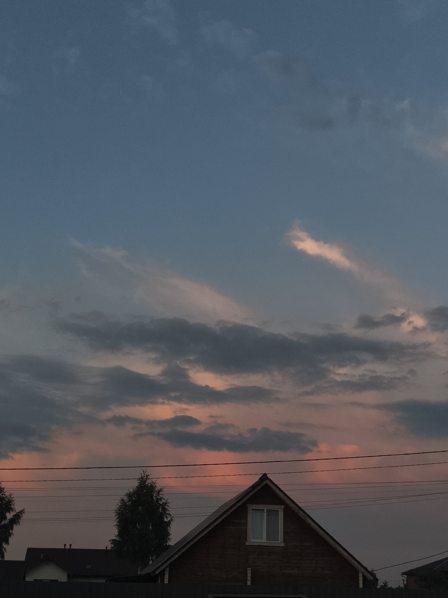 A twilight scene in Русский Вожой, Russia, features a predominantly blue sky transitioning to shades of pink and orange near the horizon due to sunset. Wispy clouds are scattered across the sky, with some illuminated by the setting sun. In the foreground, the roofline of a wooden house with a white-framed window is visible, along with power lines stretching horizontally across the frame. Silhouetted trees and other structures are present in the lower portion of the image, suggesting a suburban or rural residential area.