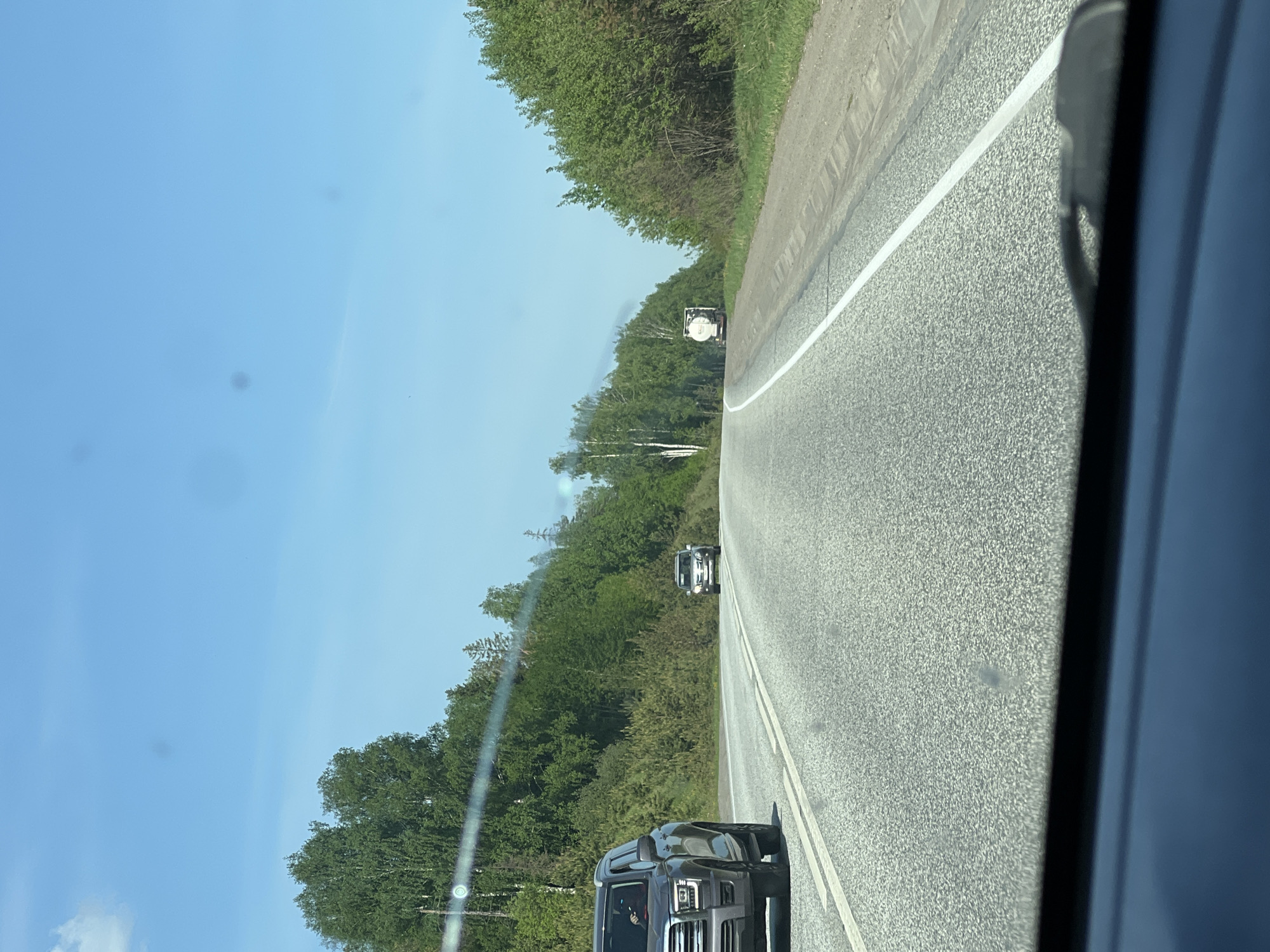 A roadside view, likely from within a moving vehicle on a road with a dashed white center line. In the distance, a white truck or trailer can be seen. Further up the road is another vehicle, likely a car or SUV. In the foreground, a dark colored pickup truck is present in the opposite lane. The environment includes trees and blue sky. The location may be in the city of Банное, Russia.