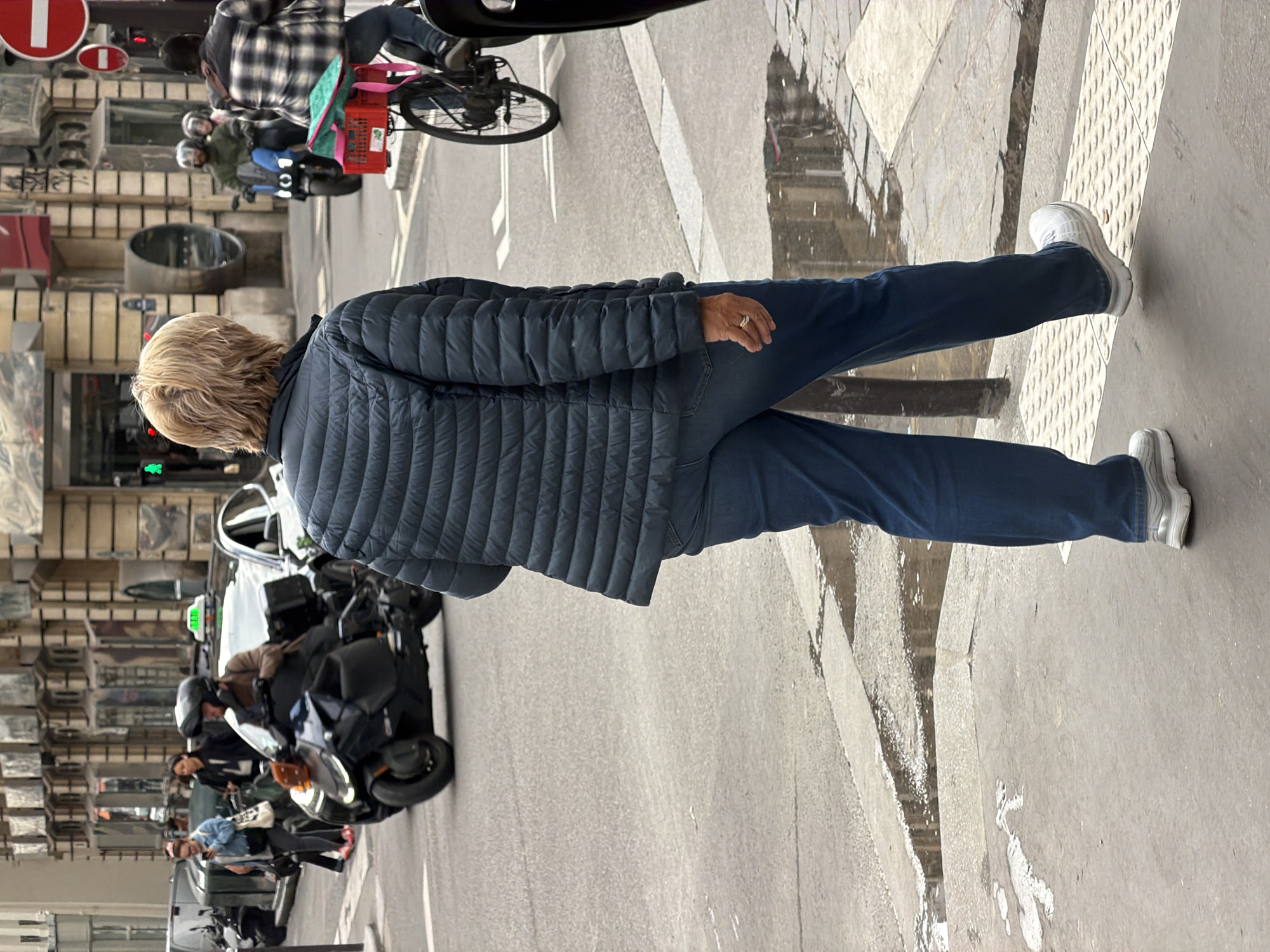 A woman with short blond hair, wearing a blue quilted jacket and blue jeans, walks across a sidewalk in Paris, France. She wears white sneakers. The street scene includes motorcycles, a bicycle with a delivery box, and parked cars, with reflections visible on the wet pavement. Buildings line the street, with signs indicating a city location.